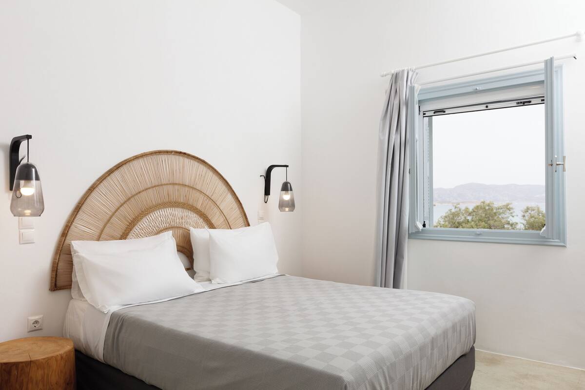 Een slaapkamer met een bed, twee wandlampen, een raam met gordijnen en uitzicht op het landschap.
