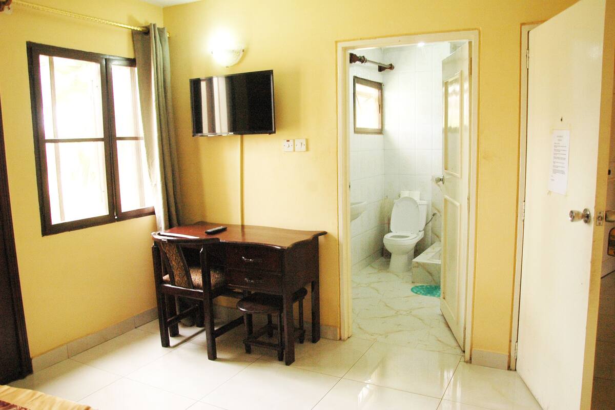 A room with a desk, chair, and a television. There is a window with curtains, a door leading to a bathroom, and a tiled floor.