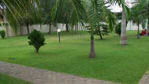 A well-maintained grassy area with a paved walkway, palm trees, and a small bush. - Pekan Hotel (Accra)