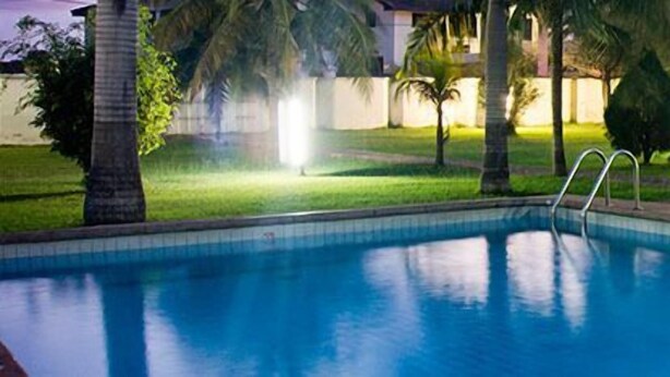A swimming pool with a thatched umbrella, surrounded by palm trees and a well-maintained lawn. - Pekan Hotel (Accra)