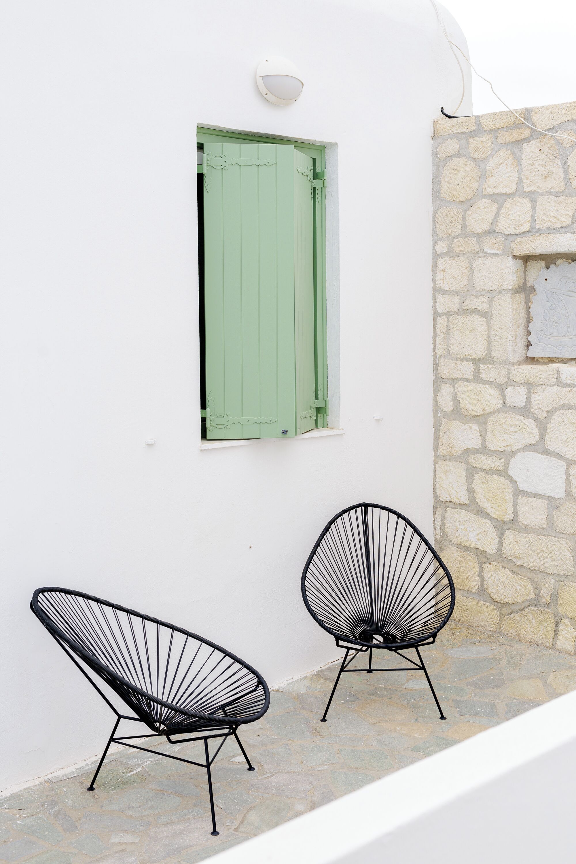 Deux chaises au design minimaliste, avec une structure en fil de fer noir, sont placées devant un mur blanc, avec un volet de fenêtre vert et un mur en pierre.