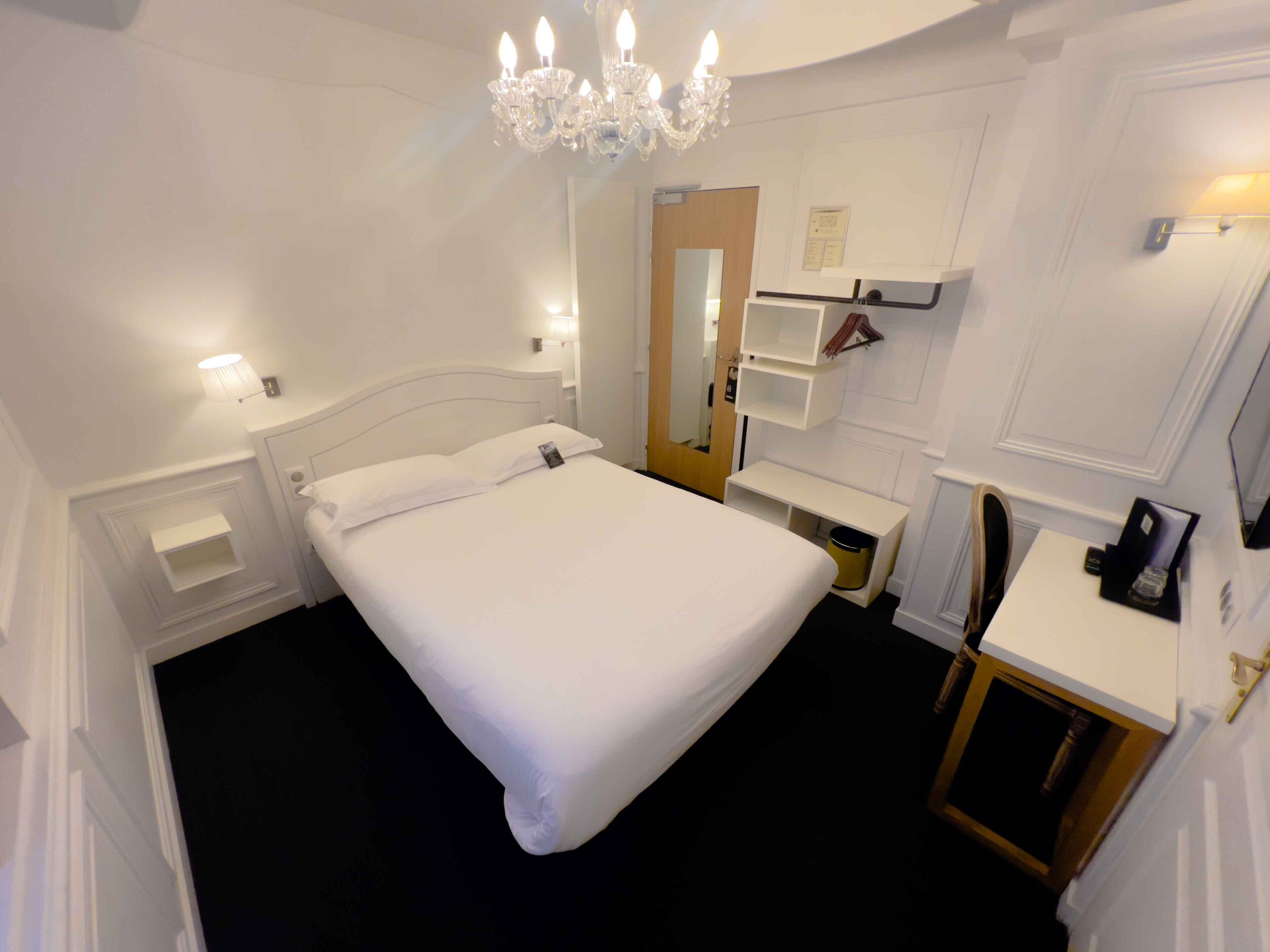 Una camera d'hotel con un letto grande, un comodino, uno specchio, un lampadario e una piccola scrivania con una lampada.