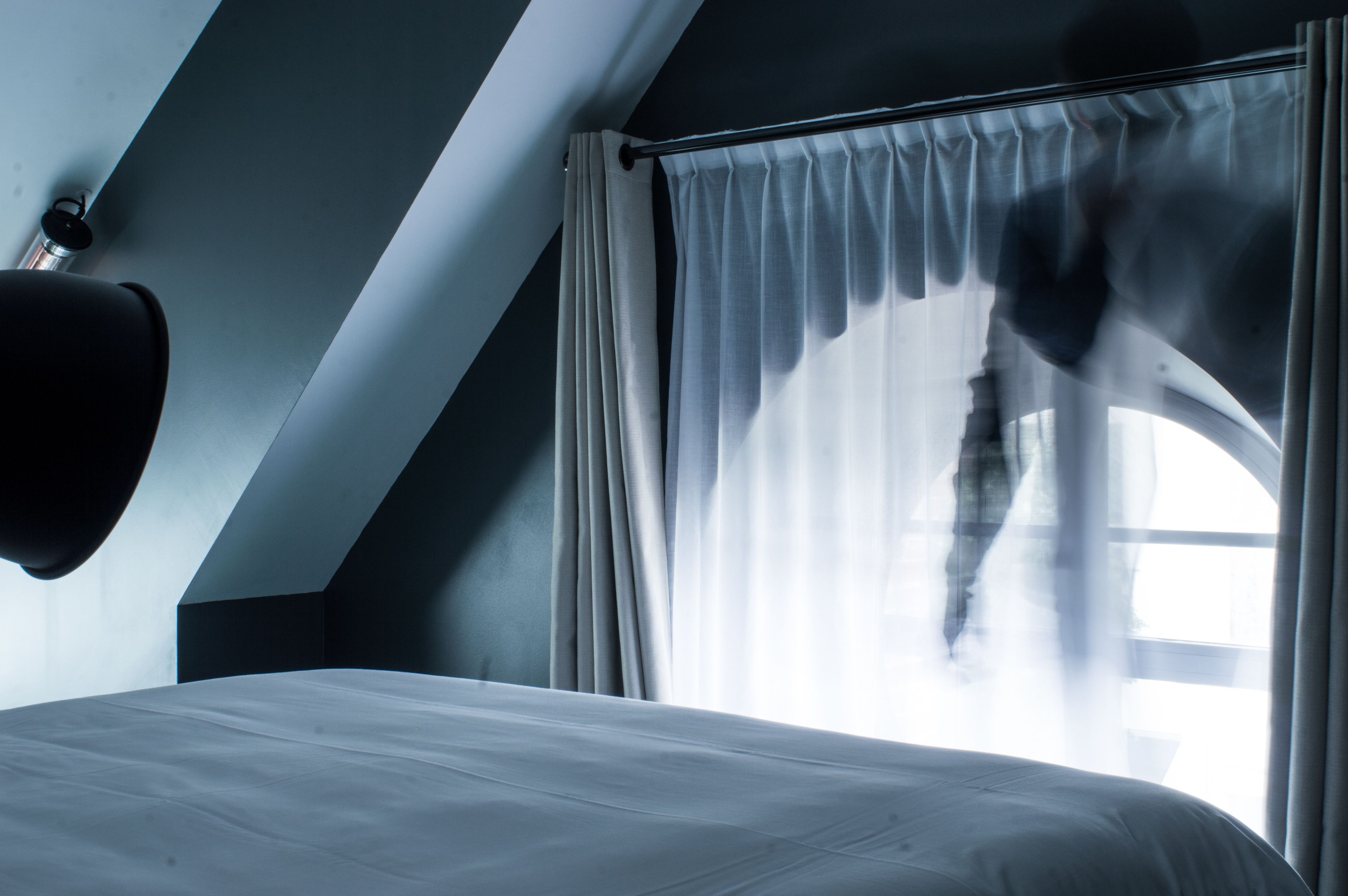 A bedroom with a bed, a window with sheer curtains, and a black wall-mounted lamp.