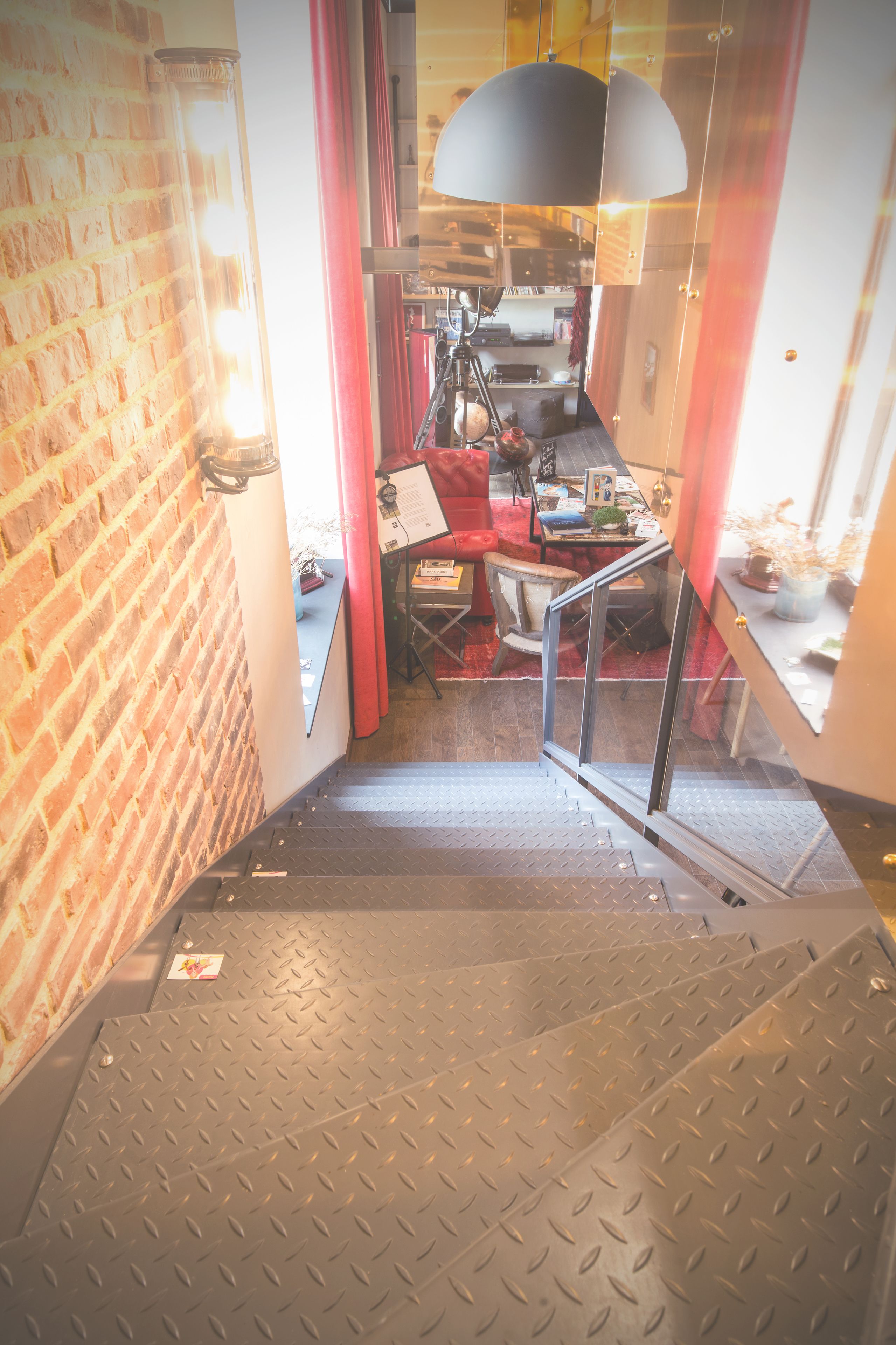 Eine Treppe mit Metallboden führt zu einem Raum mit Backsteinwand, einem roten Stuhl und einem Tisch mit verschiedenen Gegenständen.
