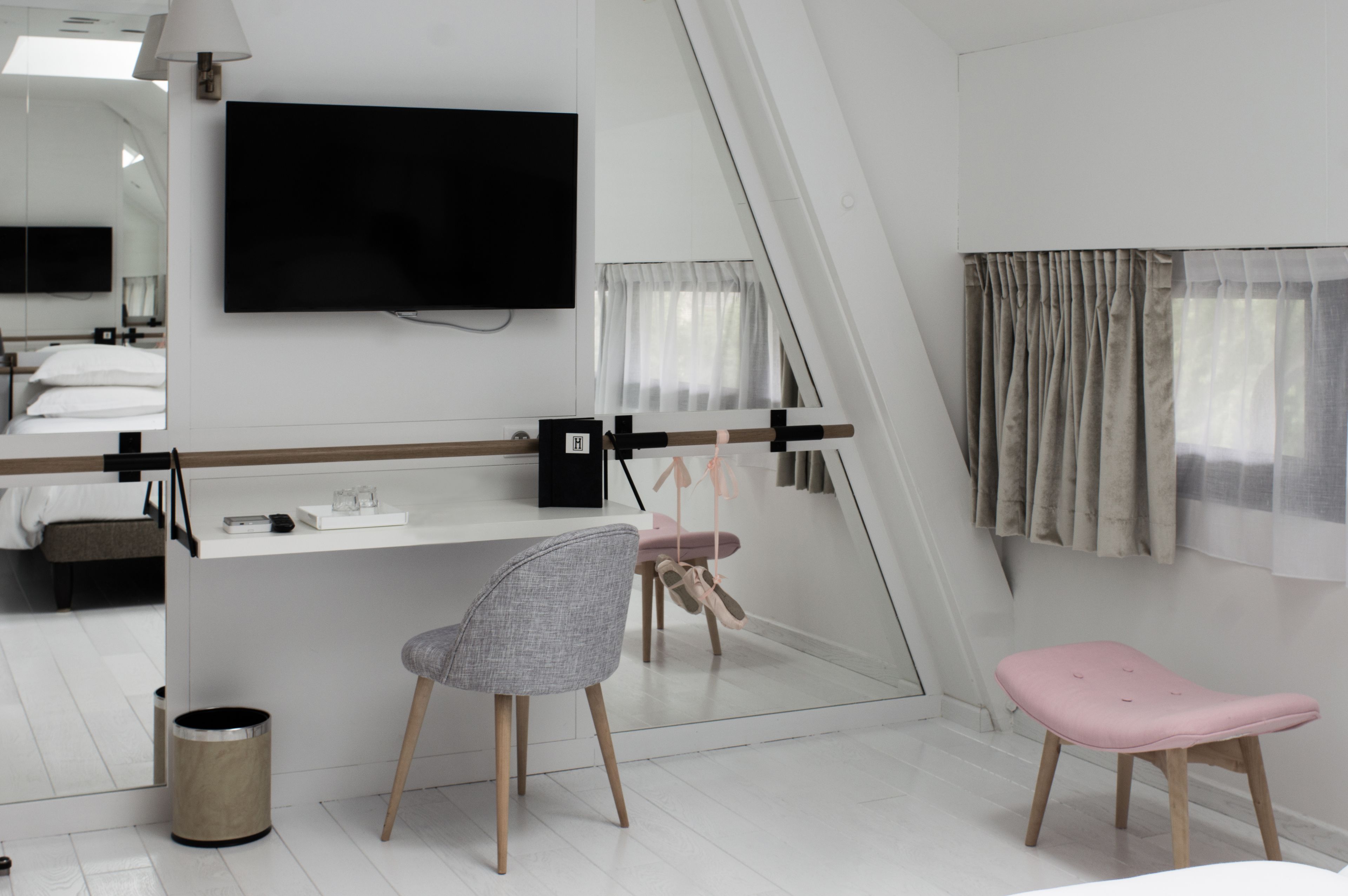 Habitación de hotel moderna con un espejo grande, un televisor de pantalla plana, un escritorio pequeño, una silla, un taburete rosa y una ventana con cortinas.