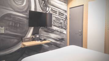 A hotel room with a bed, a flat-screen TV, a desk with a radio, and a wall mural.