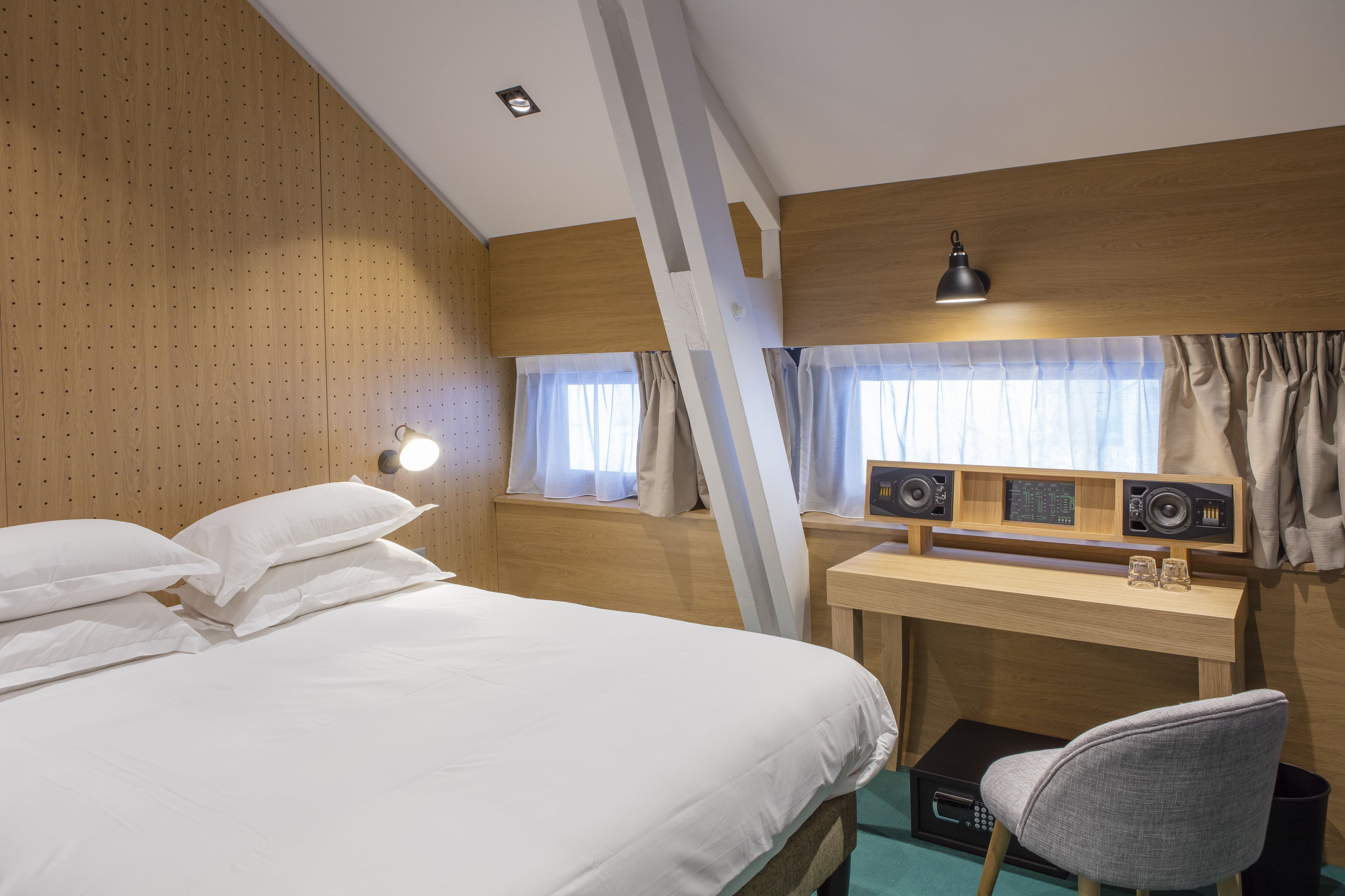 Hotellihuone, jossa on suuri sänky, työpöytä kaiuttimilla, tuoli ja ikkuna, jossa on verhot.