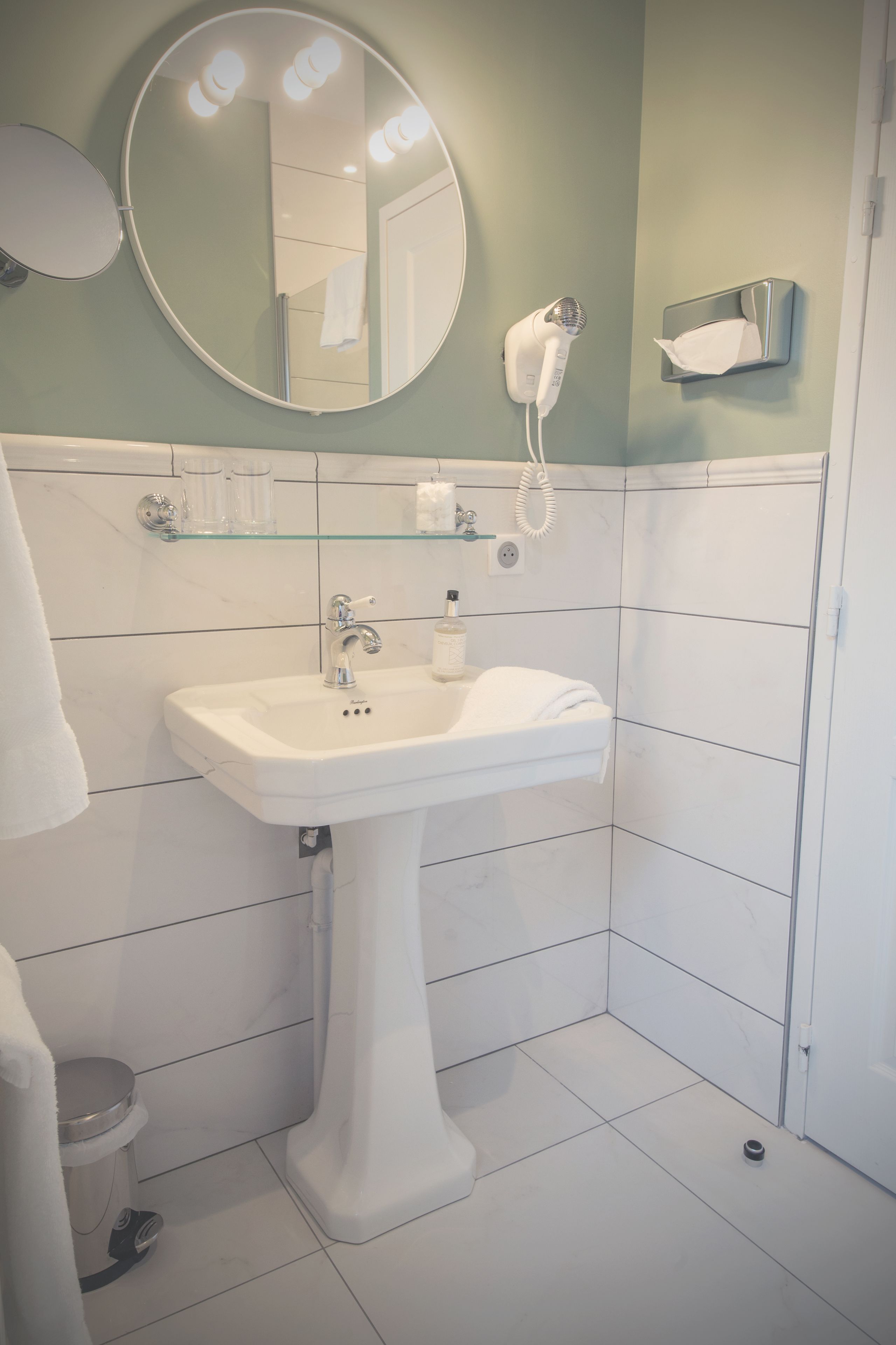 A bathroom with a pedestal sink, a round mirror, a hairdryer, and a tissue dispenser.