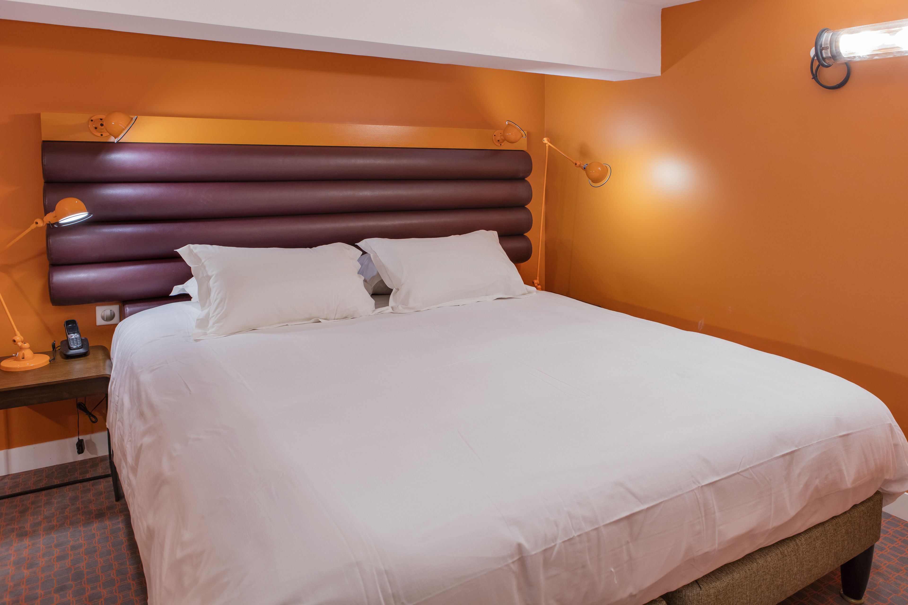 A neatly made bed with white linens, a wooden nightstand with a lamp, and a telephone.
