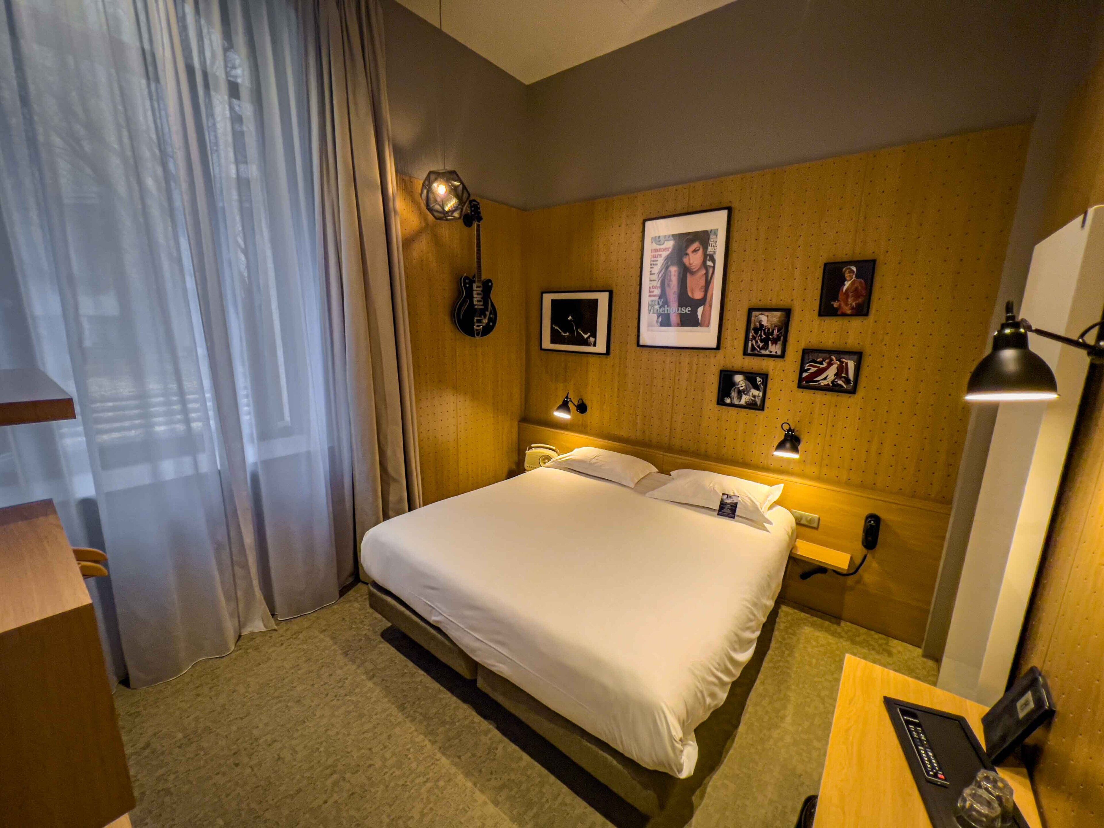 A hotel room with a large bed, a desk with a lamp, and a wall with framed pictures.