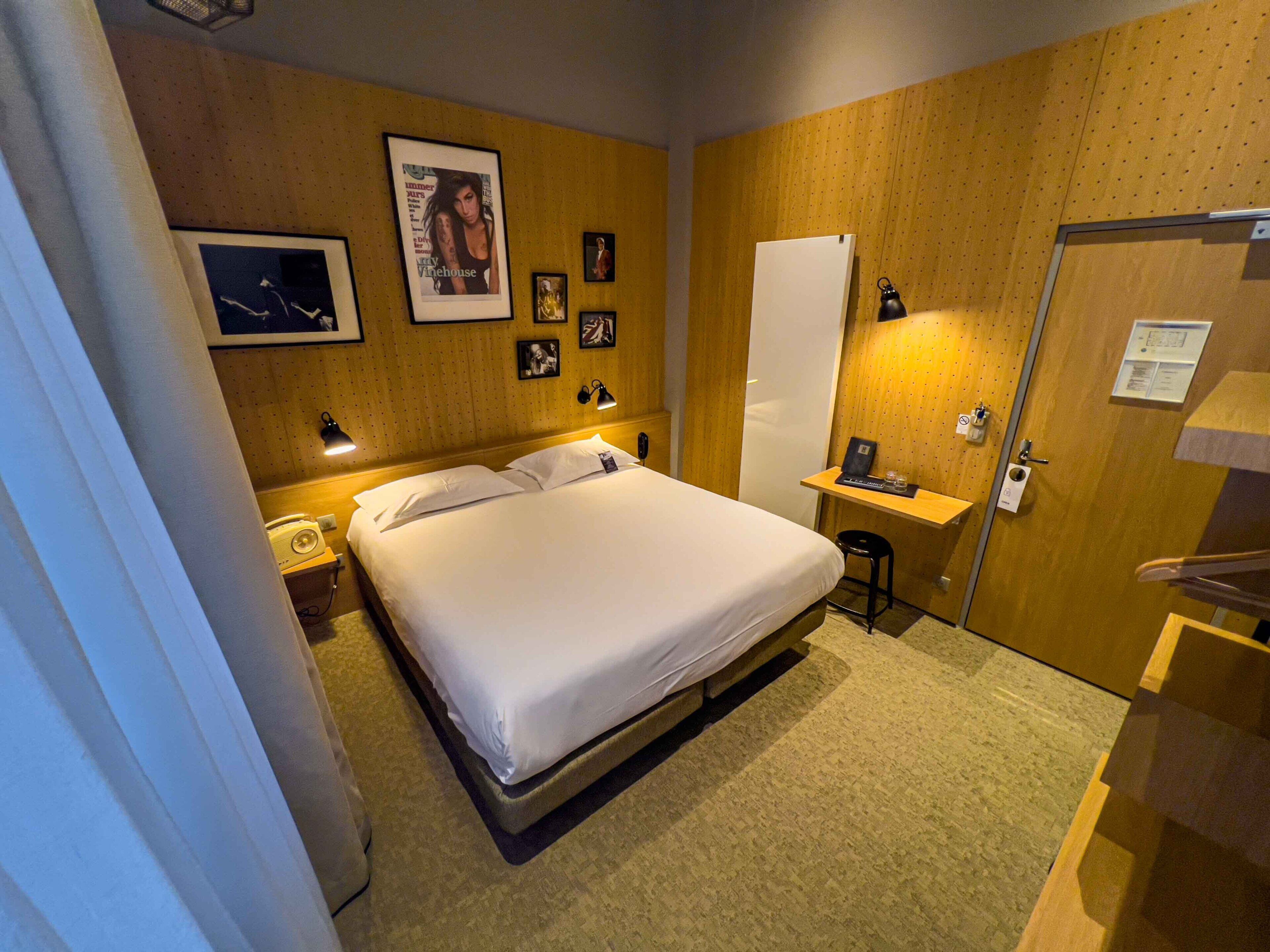 A hotel room with a bed, bedside table, lamp, and a small stool.
