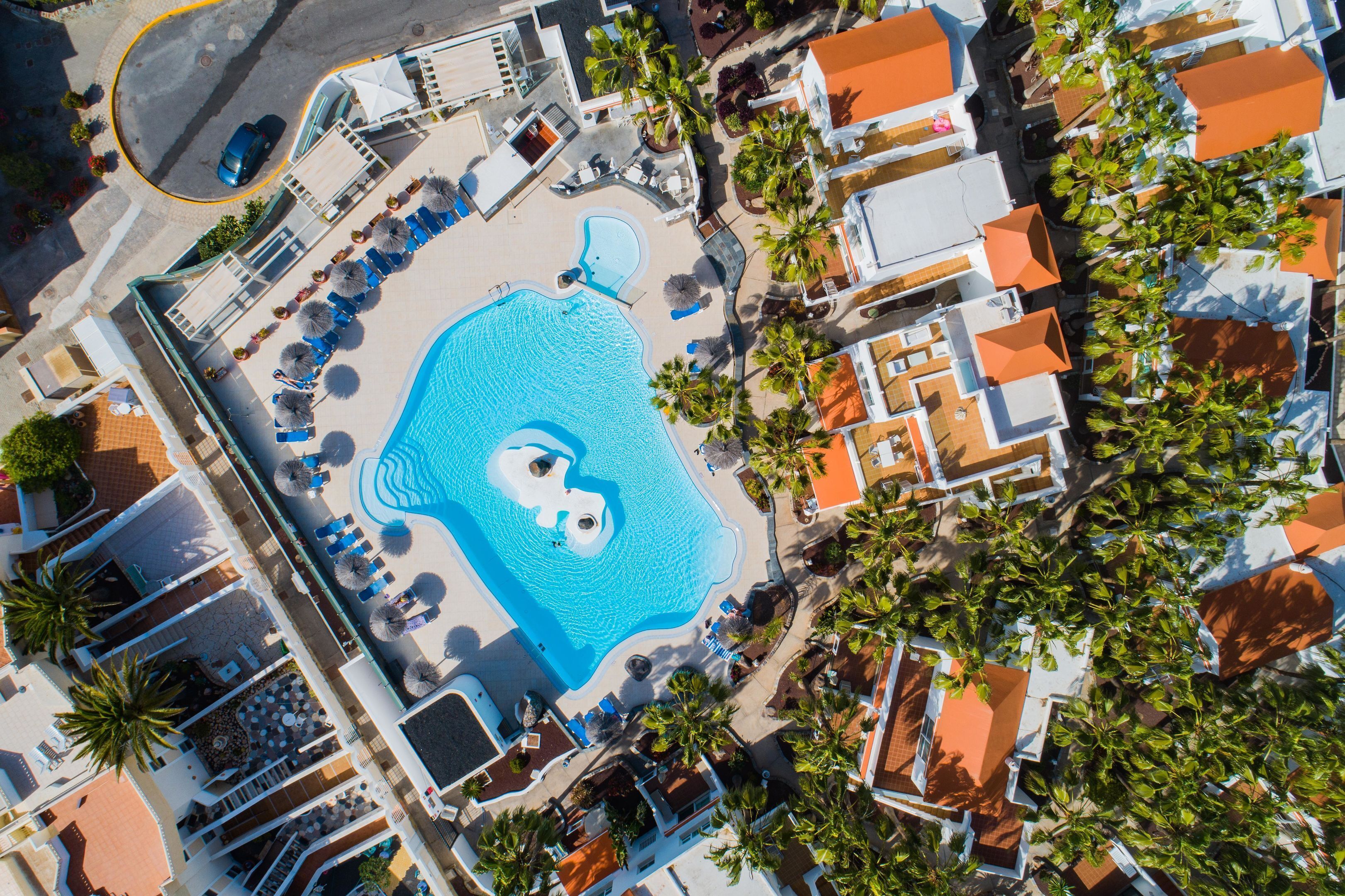 Vue aérienne d’un complexe hôtelier doté d’une piscine centrale, de plusieurs espaces de détente et de palmiers.