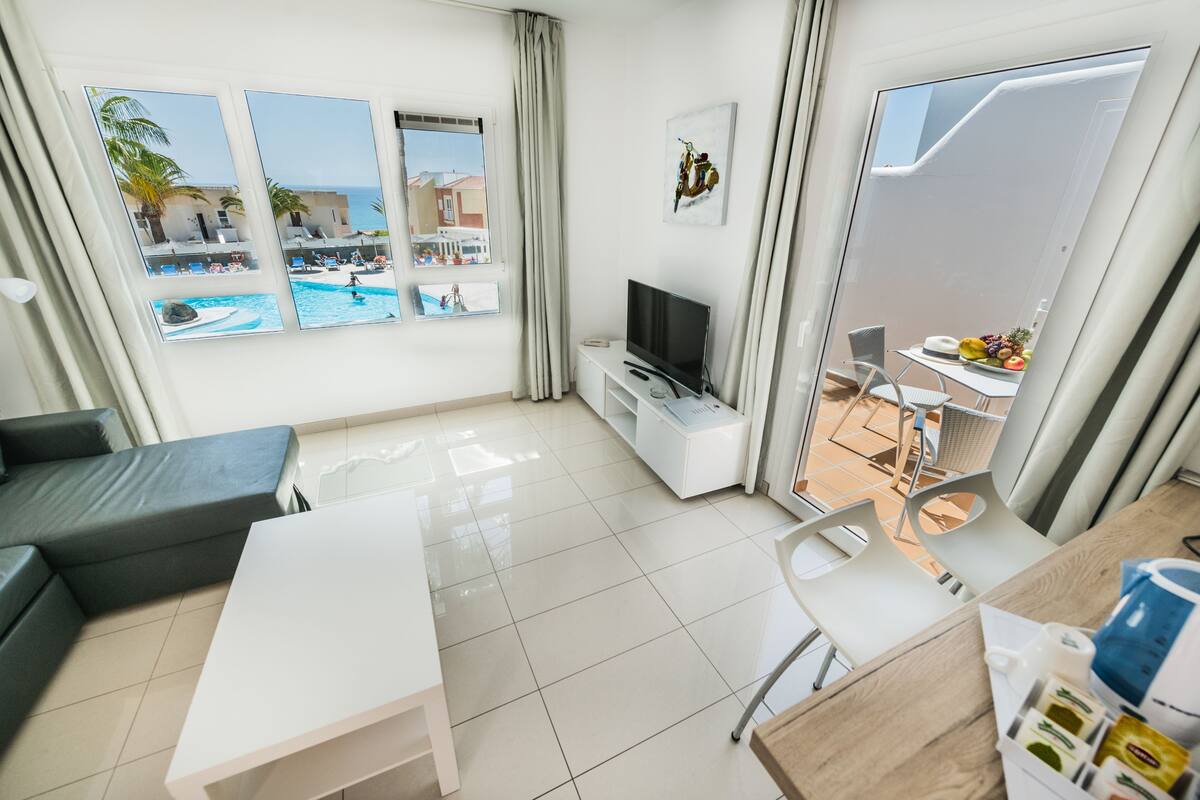 A modern hotel room with a large window overlooking a pool and a view of the sea. There is a flat-screen TV, a dining table, and a small kitchen area with a coffee maker.