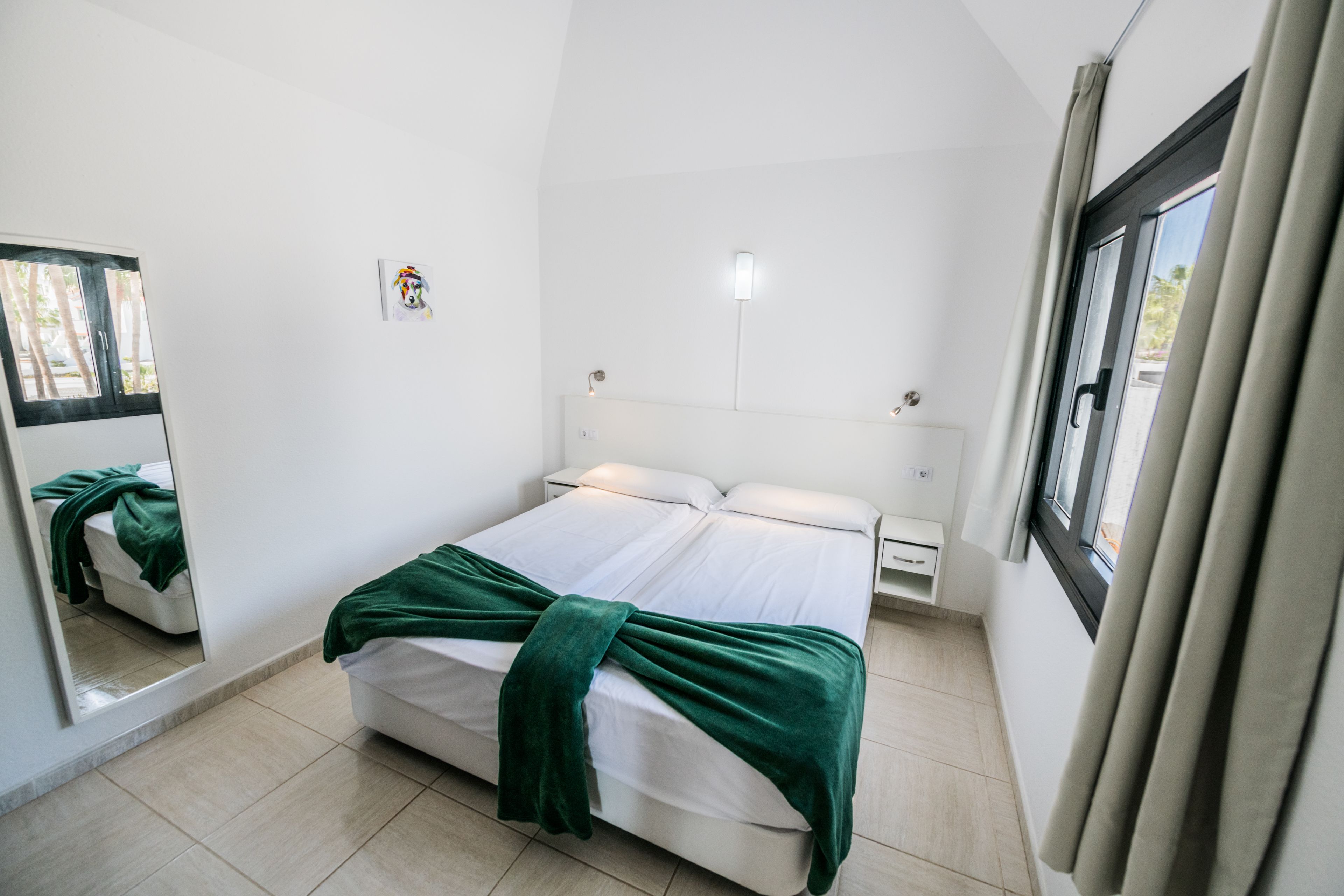 Un dormitorio con cama, mesita de noche, lámpara de noche, ventana con cortinas y espejo adosado a la pared.