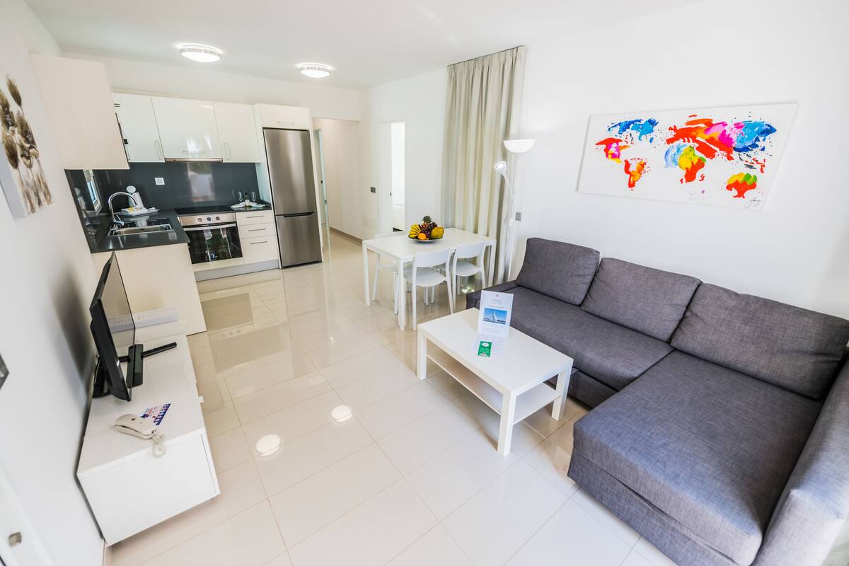 A modern living room with a grey sofa, a white coffee table, and a dining area with a white table and chairs. A kitchen with white cabinets and a stainless steel refrigerator is visible in the background.