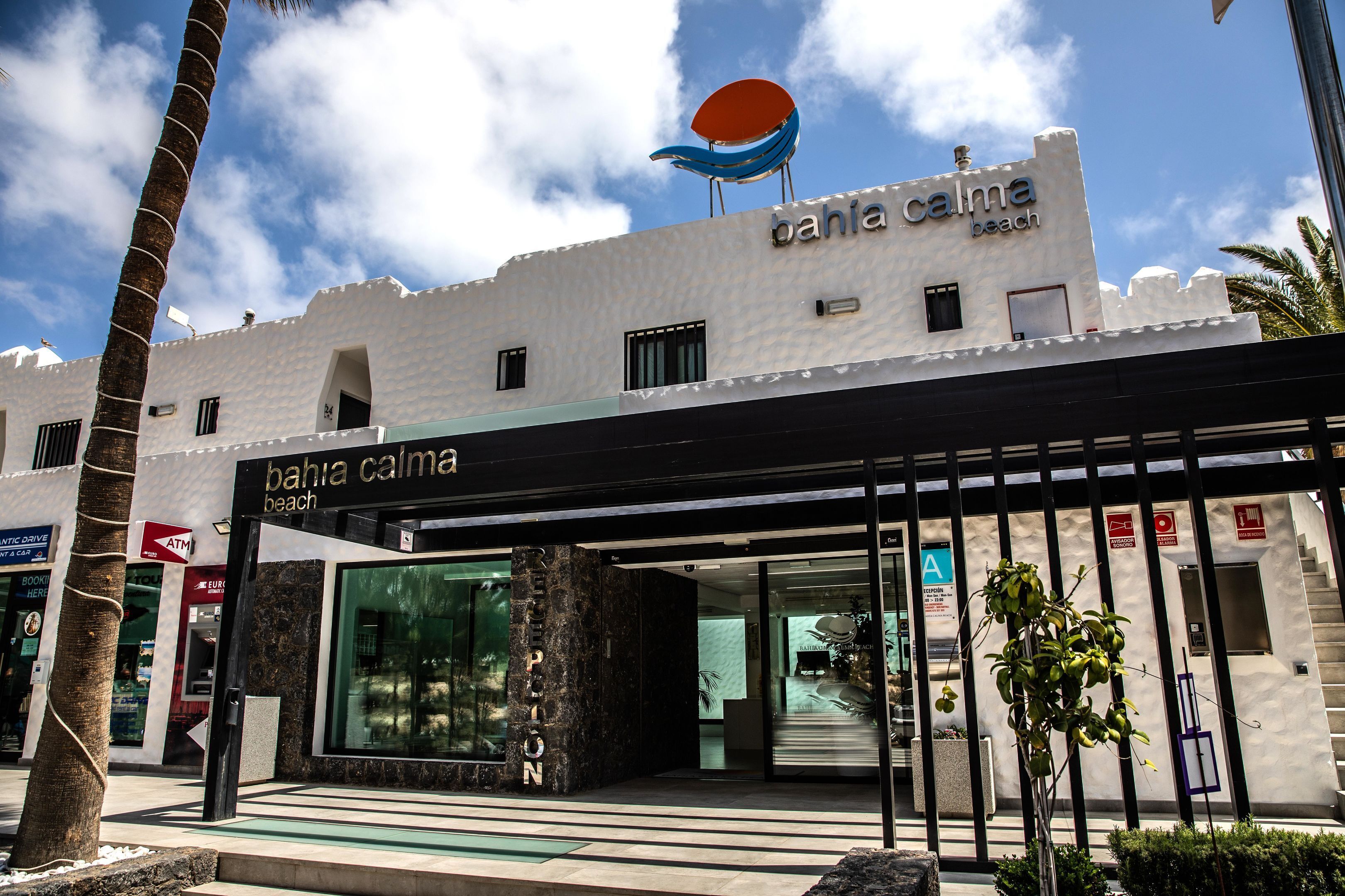 Een hotel genaamd 'Bahia Calma Beach' met een moderne gevel, een grote glazen entree en een opvallend blauw-oranje bord op het dak.