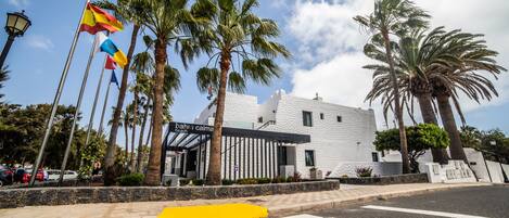 A modern building with a white facade, palm trees, and flags.