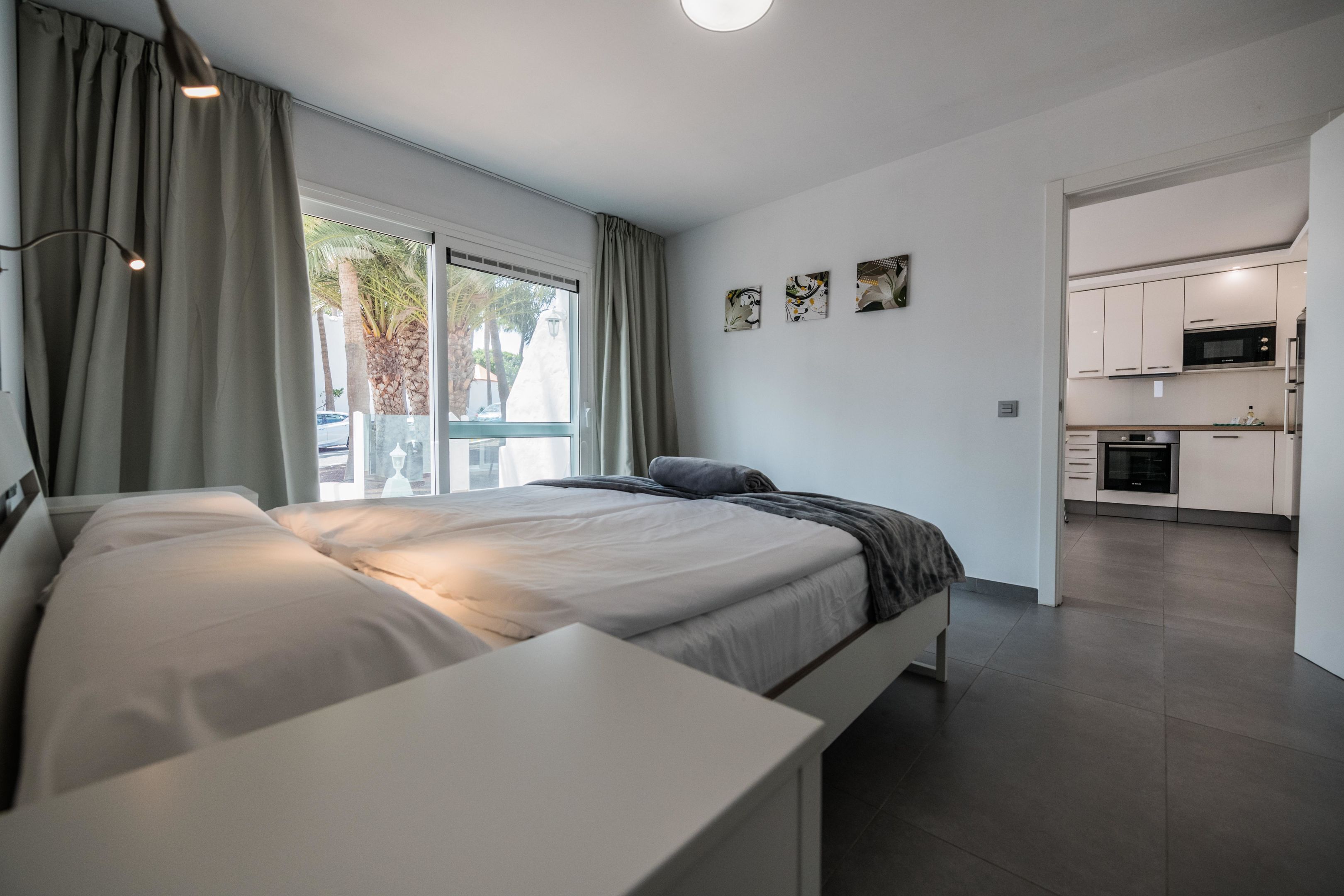 A modern bedroom with a large bed, a bedside table, and a view of the kitchen through an open door.