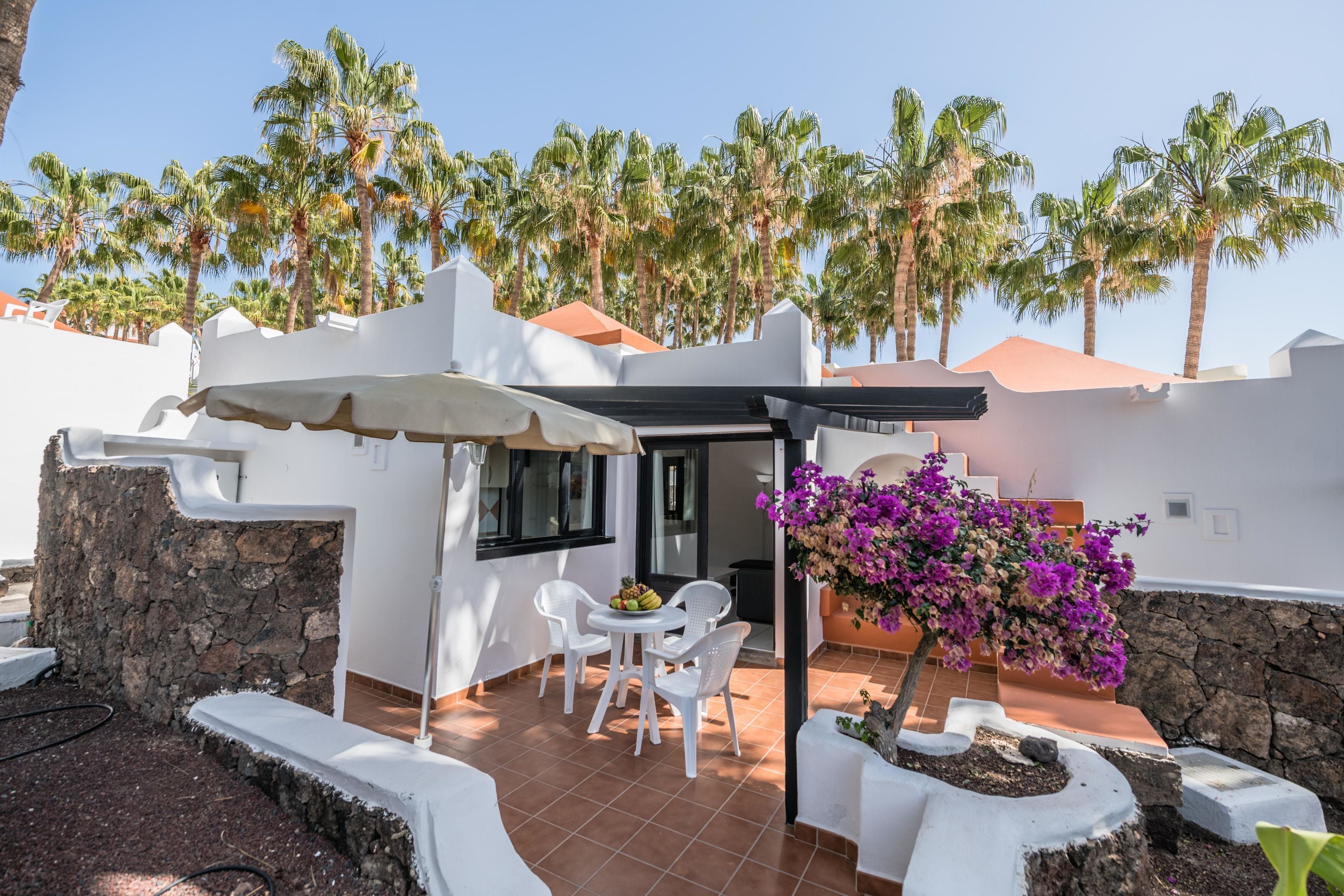 A patio area with white chairs, a small table with fruit, and a purple flowering plant.