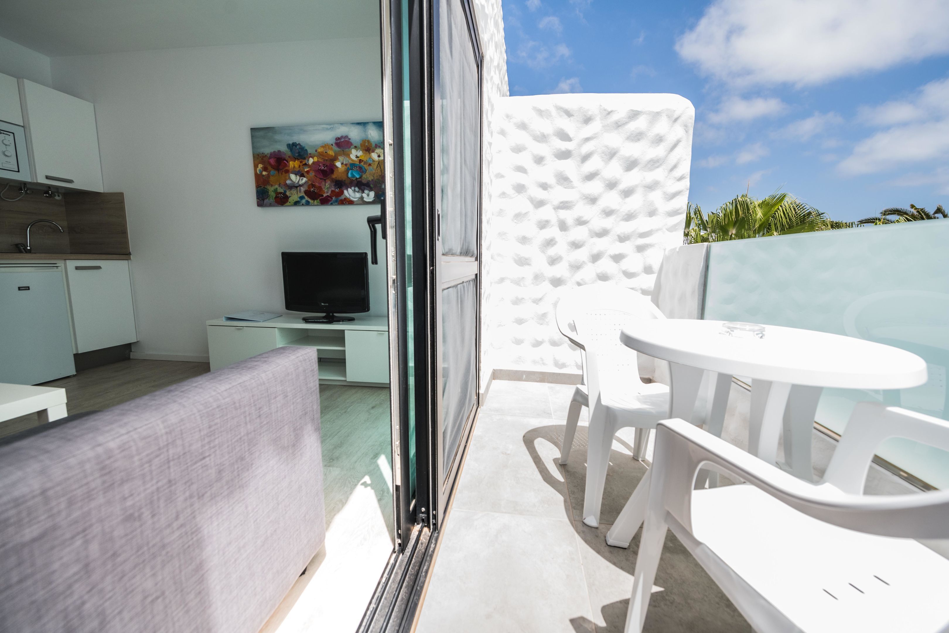A modern, well-lit room with a sofa, a TV, a small table, and a balcony with white chairs and a table.