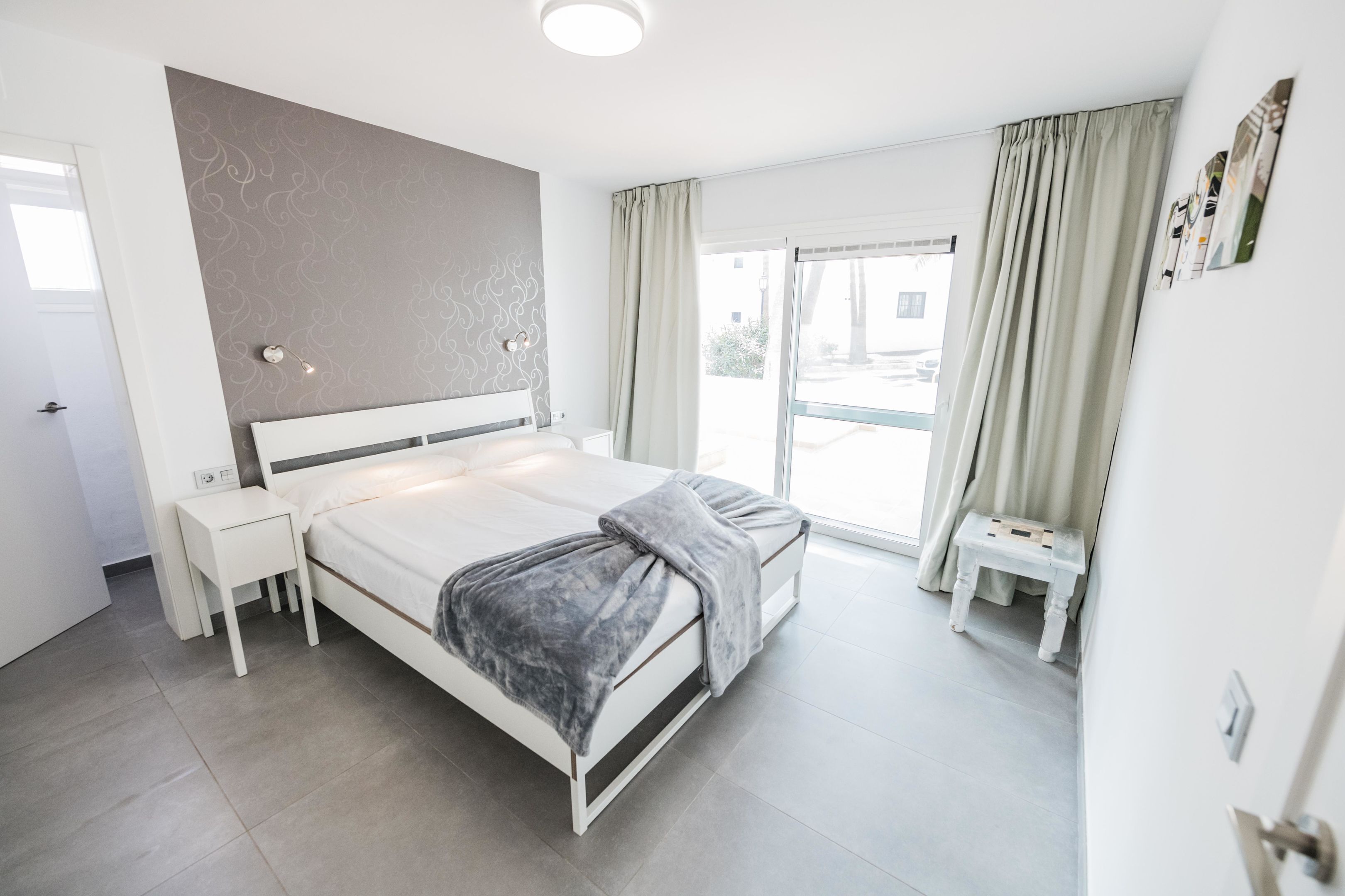 A modern bedroom with a bed, bedside tables, a small stool, and a large window with curtains.