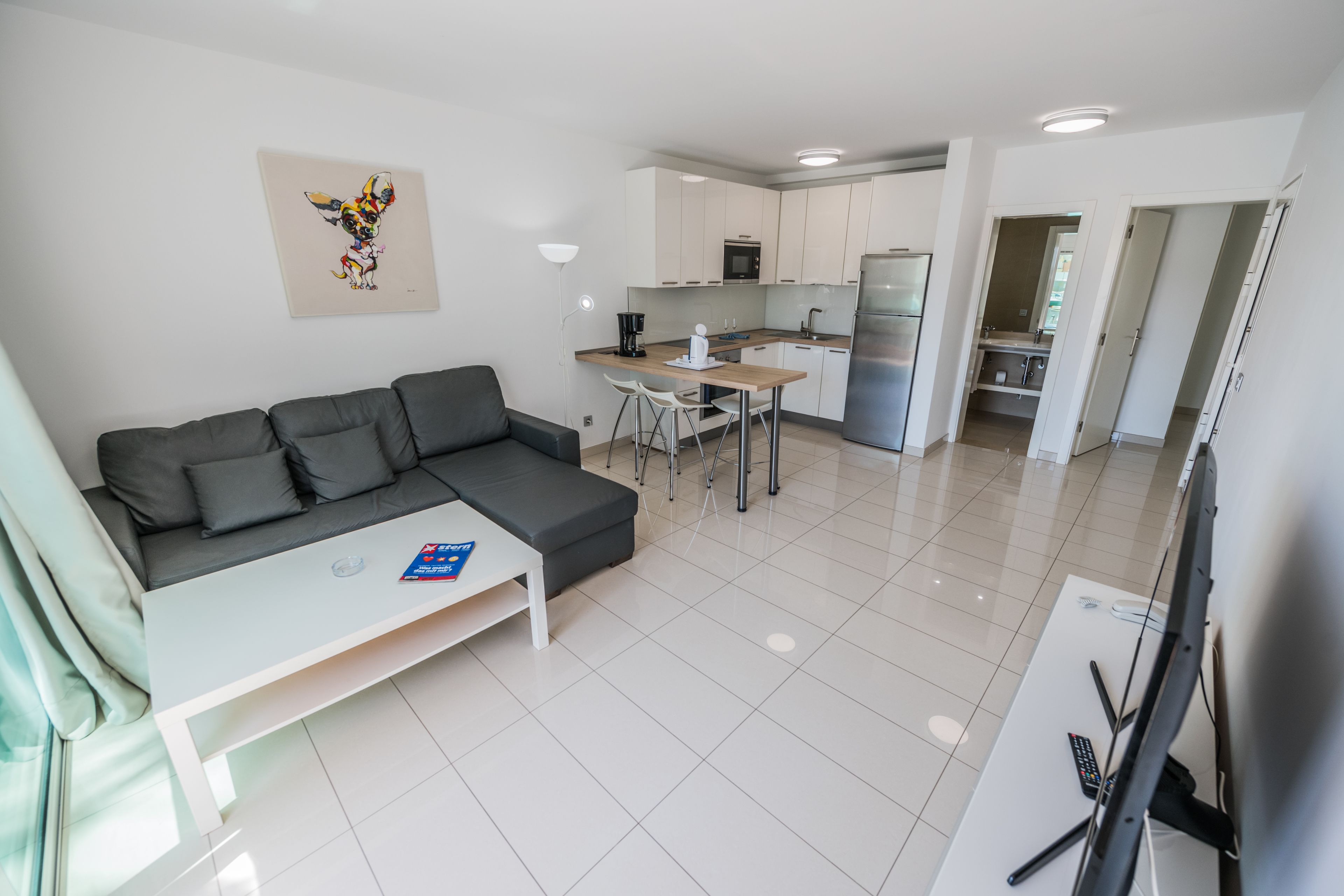 A modern living room with a grey sofa, a small white table, and a TV. A kitchen with white cabinets and stainless steel appliances is visible in the background.