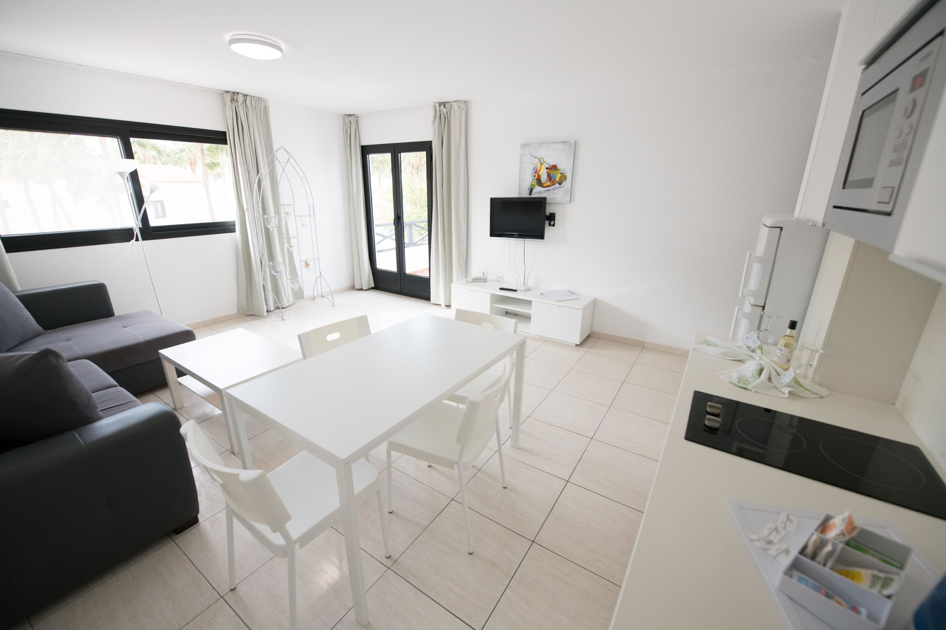 Une pièce moderne et bien éclairée, avec une table à manger blanche, des chaises, un canapé, un téléviseur à écran plat fixé au mur et un coin cuisine équipé d’un four à micro-ondes et d’une cuisinière.