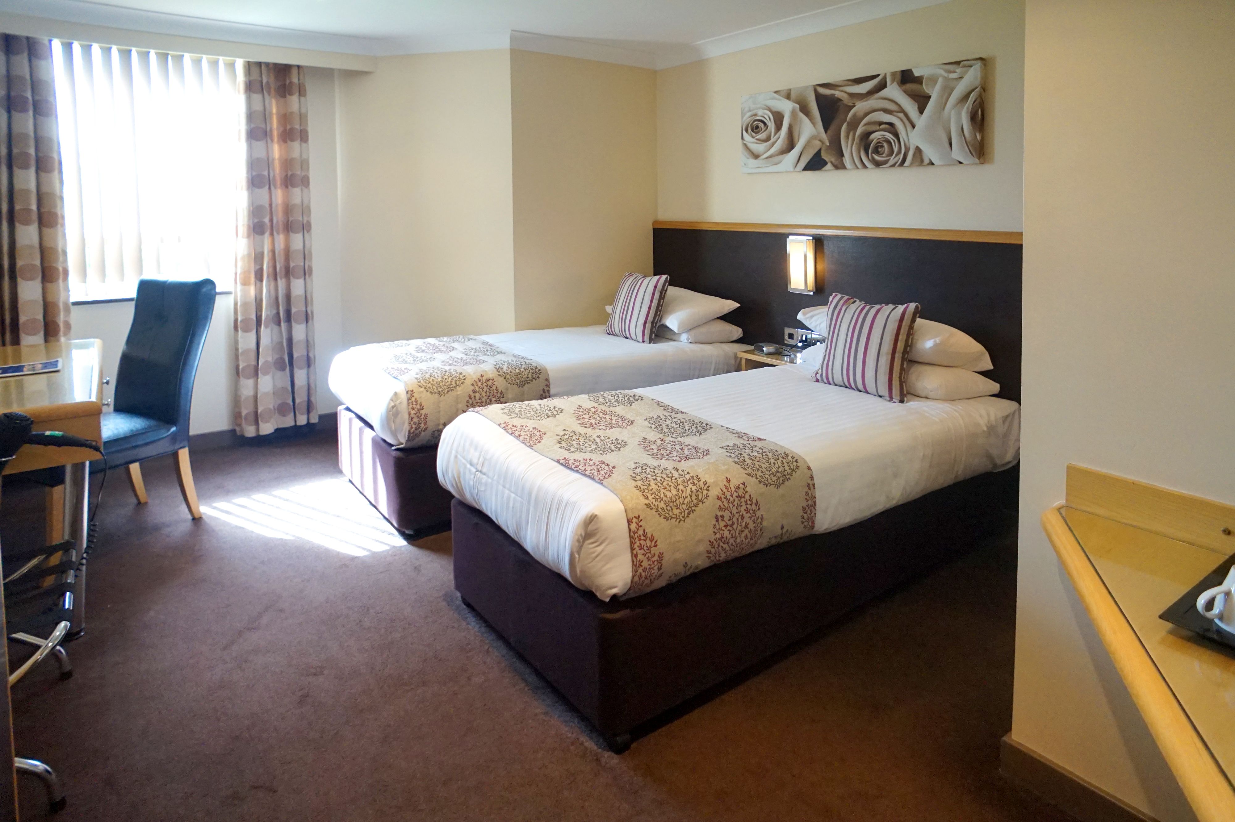 A hotel room with two beds, a desk, a chair, and a large window with curtains.