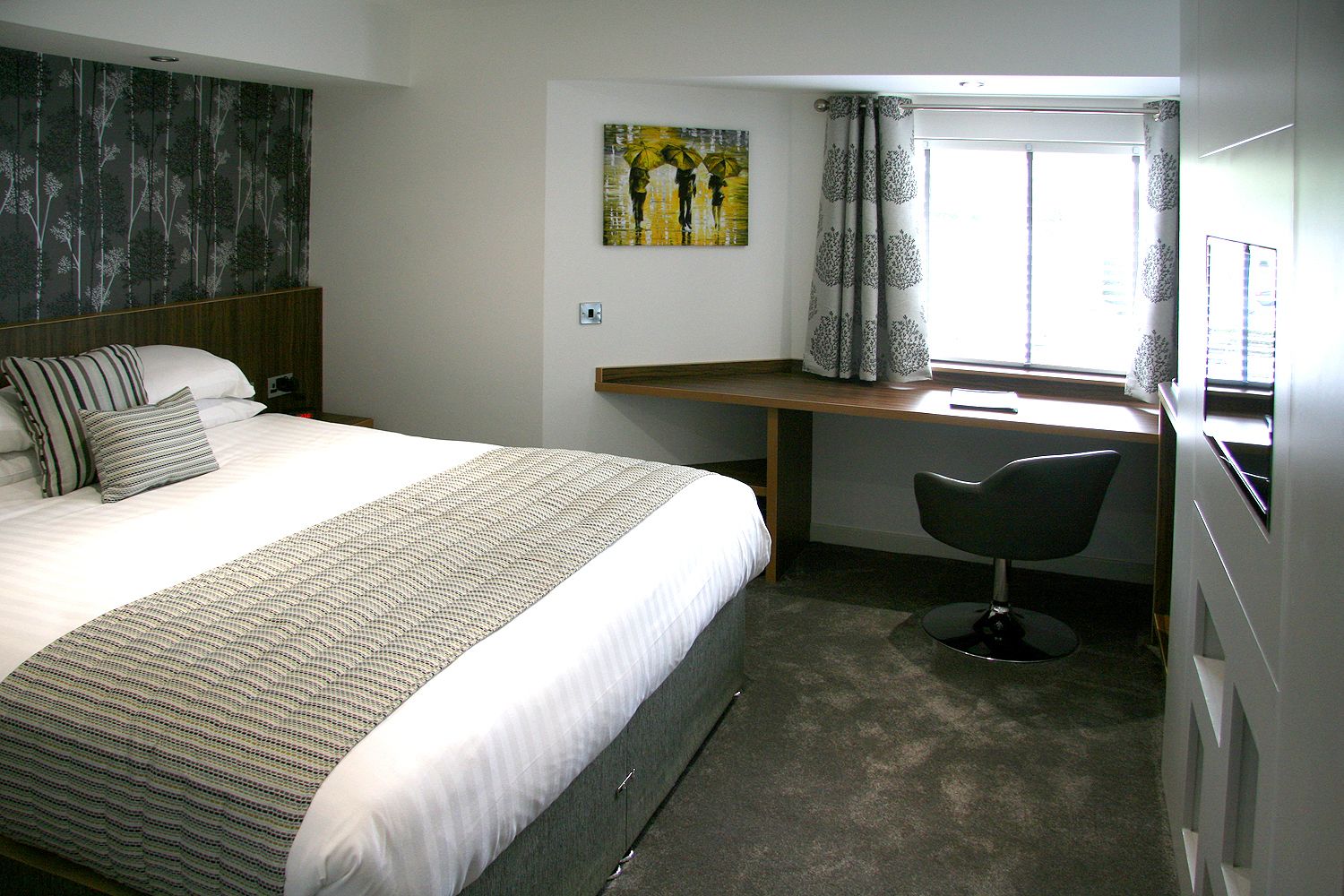 Habitación de hotel con cama, escritorio, silla, un cuadro y ventana con cortinas.