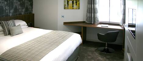 Habitación de hotel con cama, escritorio, silla, un cuadro y ventana con cortinas.