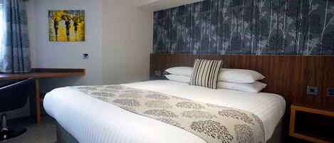 A hotel room with a bed, a desk, a chair, and a wall with a tree pattern.