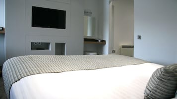 Een slaapkamer met een bed, een flatscreen televisie aan de muur, een klein plankje en een radiator.
