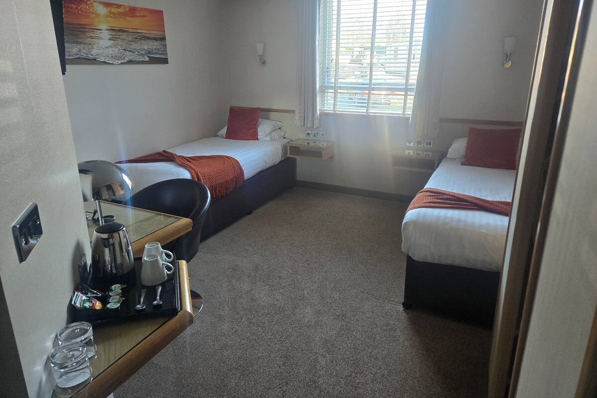 A hotel room with two single beds, a small table with a coffee maker, and a window with blinds.