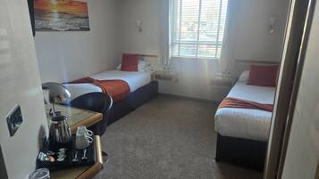 A hotel room with two single beds, a small table with a coffee maker, and a window with blinds.
