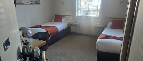 A hotel room with two single beds, a small table with a coffee maker, and a window with blinds.