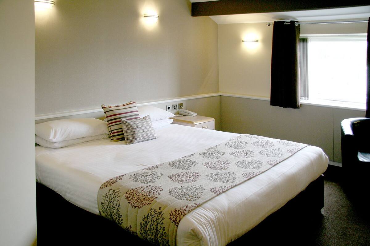 A neatly made bed with a floral-patterned bedspread, a bedside telephone, and a chair to the right.