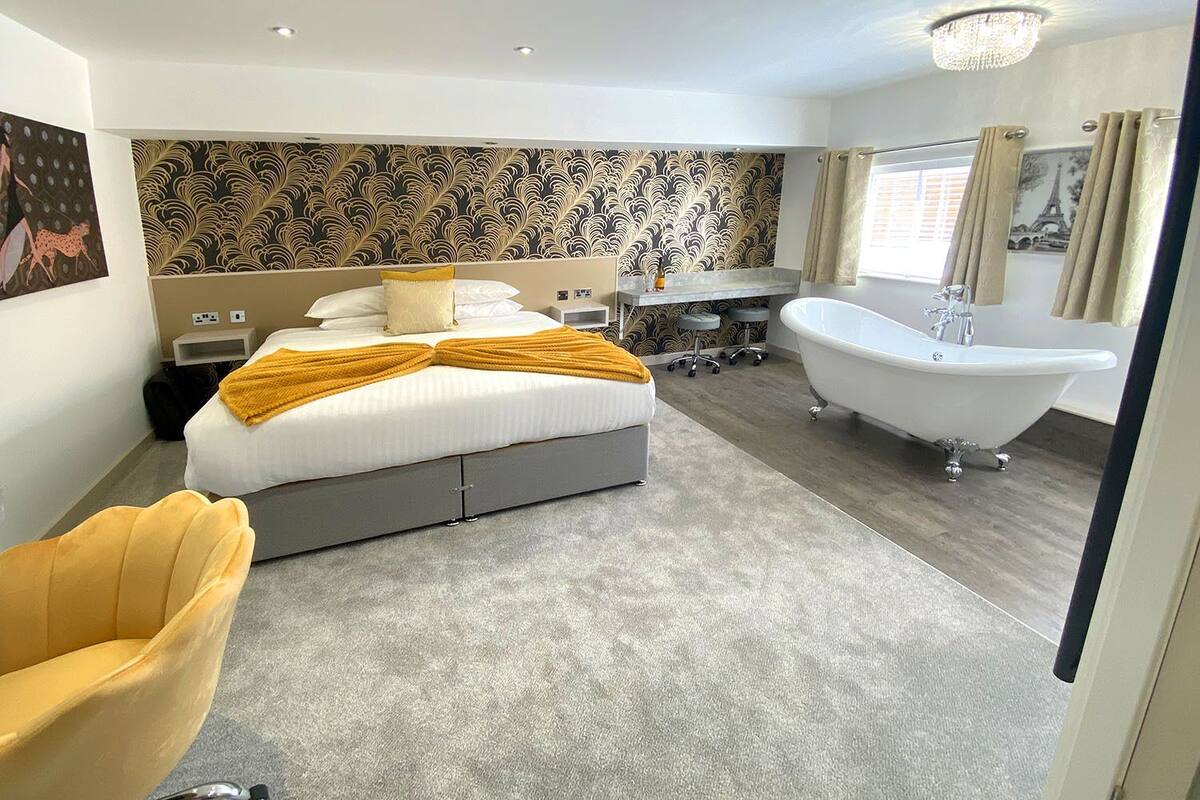 A modern bedroom with a large bed, a sitting area, a desk, and a classic bathtub.