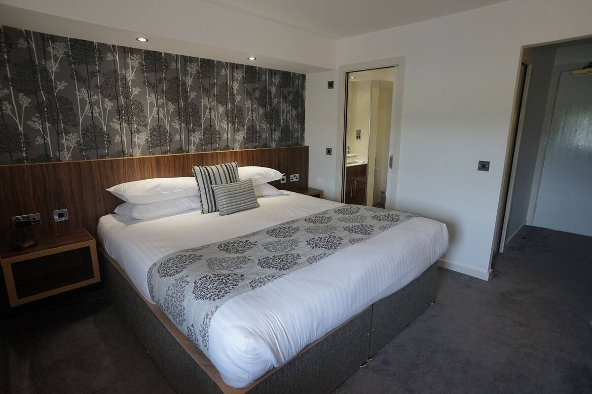A modern bedroom with a large bed, a wooden headboard, a nightstand, and a mirror.