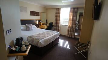 A hotel room with a bed, bedside table with coffee maker and cups, a desk with chair, and a TV mounted on the wall.