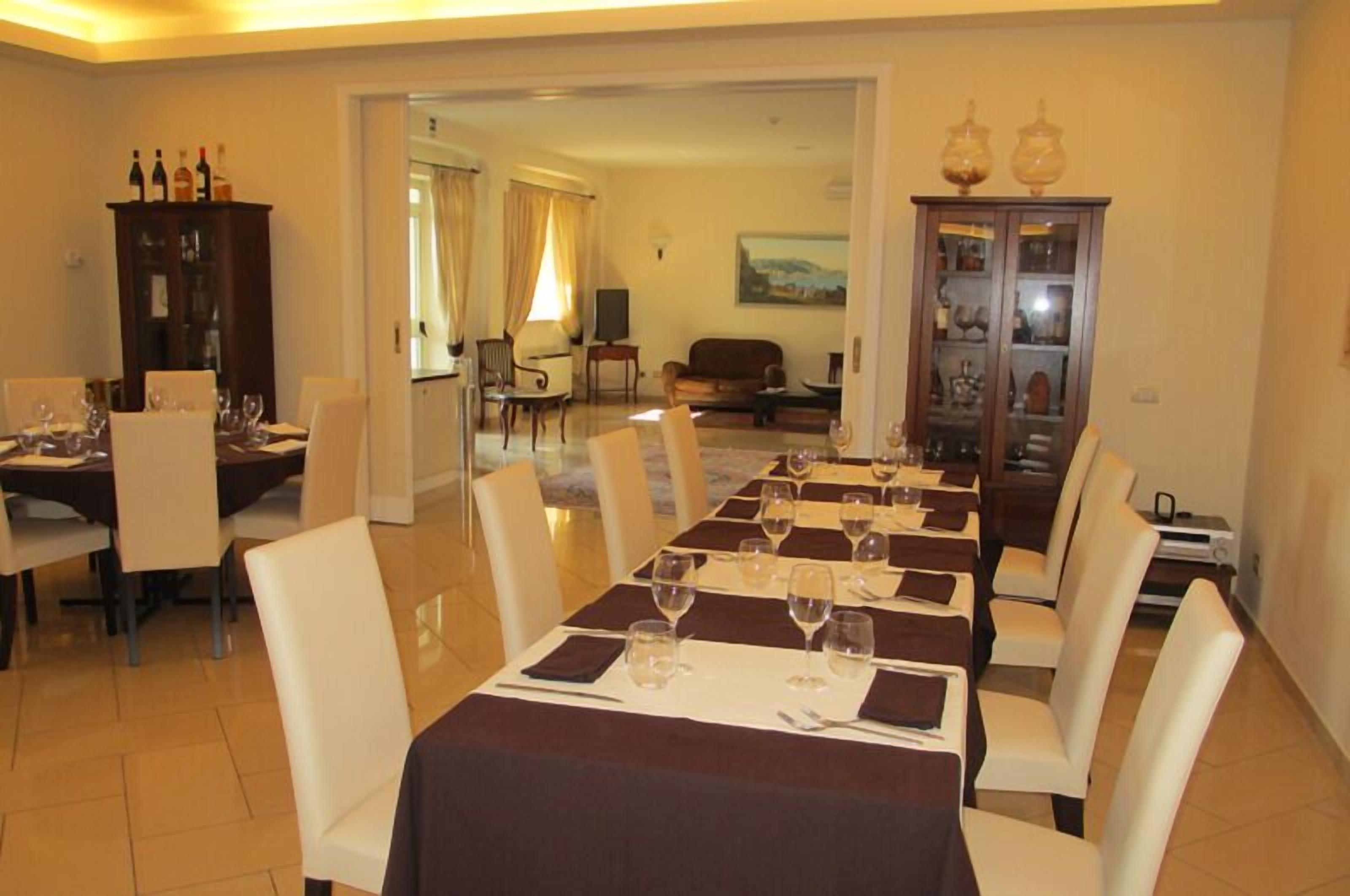 Uma sala de jantar com uma mesa comprida posta para uma refeição, rodeada de cadeiras. Há um armário com portas de vidro à direita, e uma televisão ao fundo.