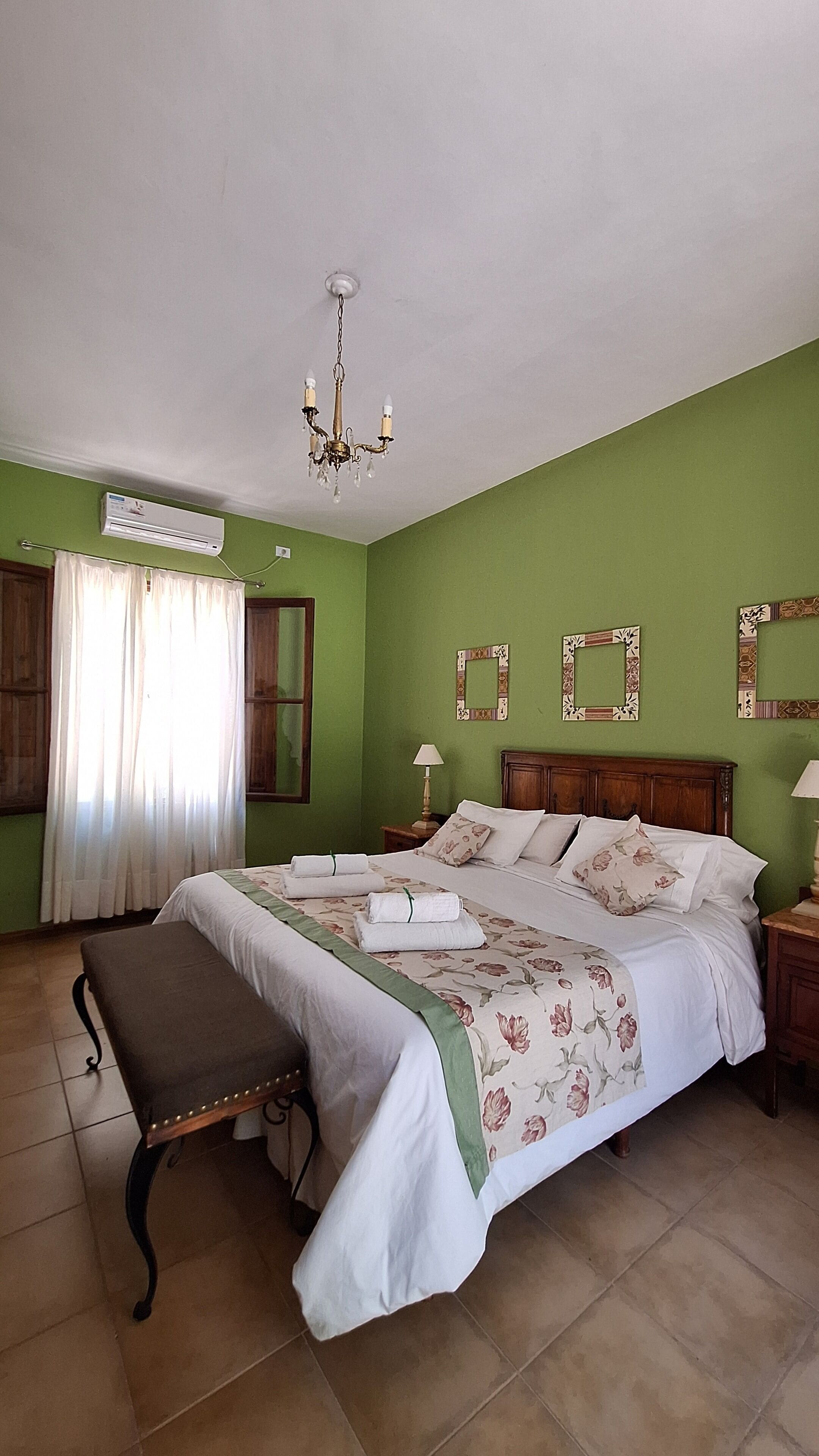 A bedroom with a bed, a bench, a chandelier, an air conditioner, and a window with curtains.