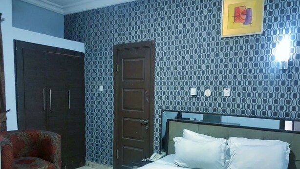 A bedroom with a bed, a chair, a wardrobe, and a patterned wall. - Prixair Pure Hotel Wuse (Abuja)