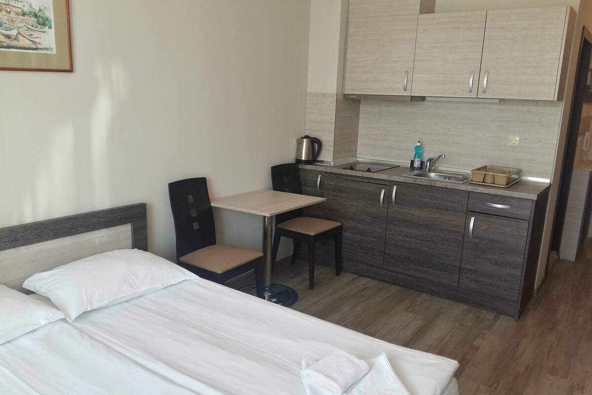 A hotel room with a bed, a small desk, a chair, a kitchenette, and a painting on the wall.
