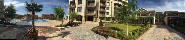 Un patio de hotel con piscina, palmeras, un banco de madera y un jardín bien cuidado.