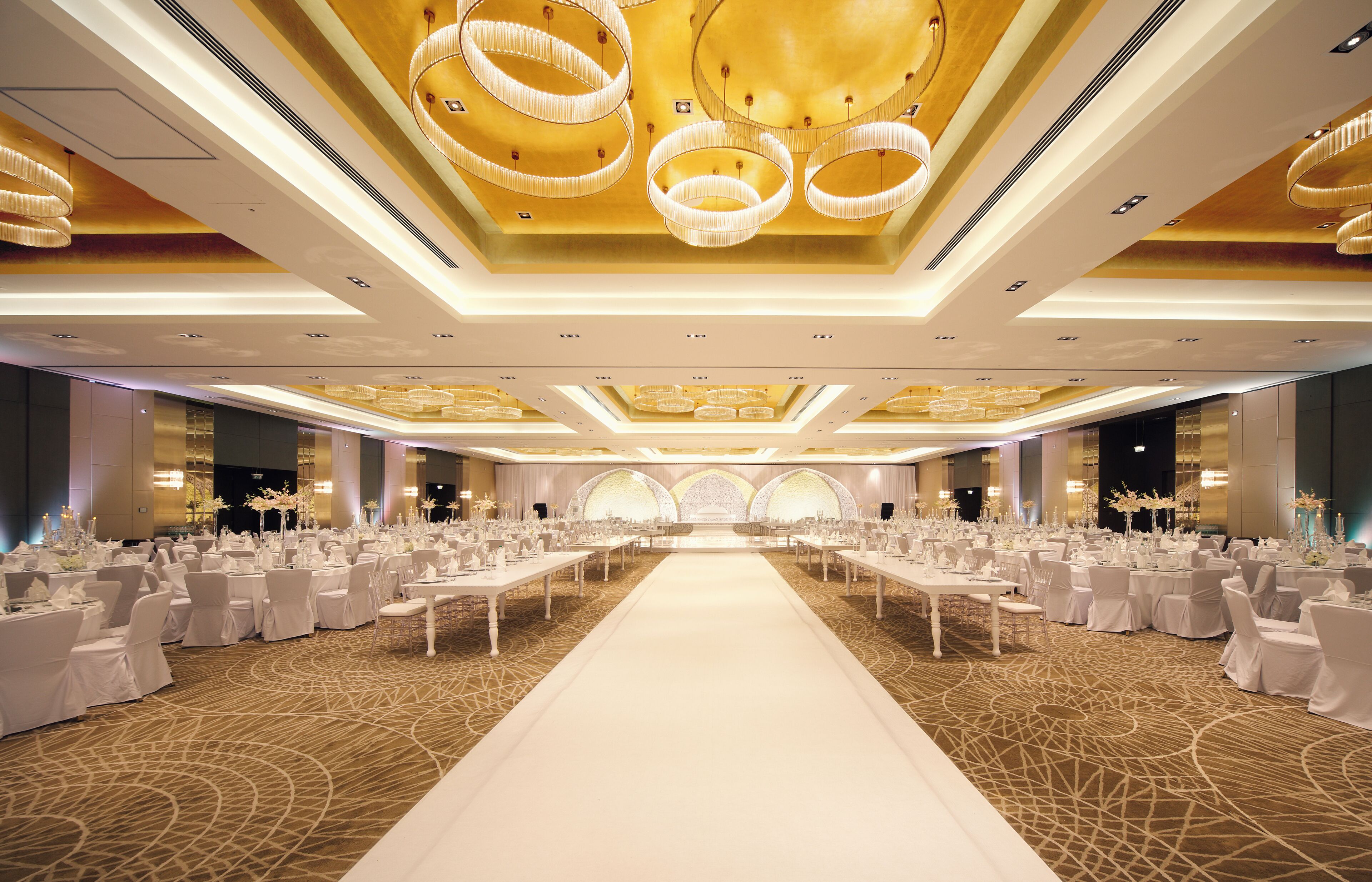 A banquet hall with a central aisle, round tables, and chandeliers.