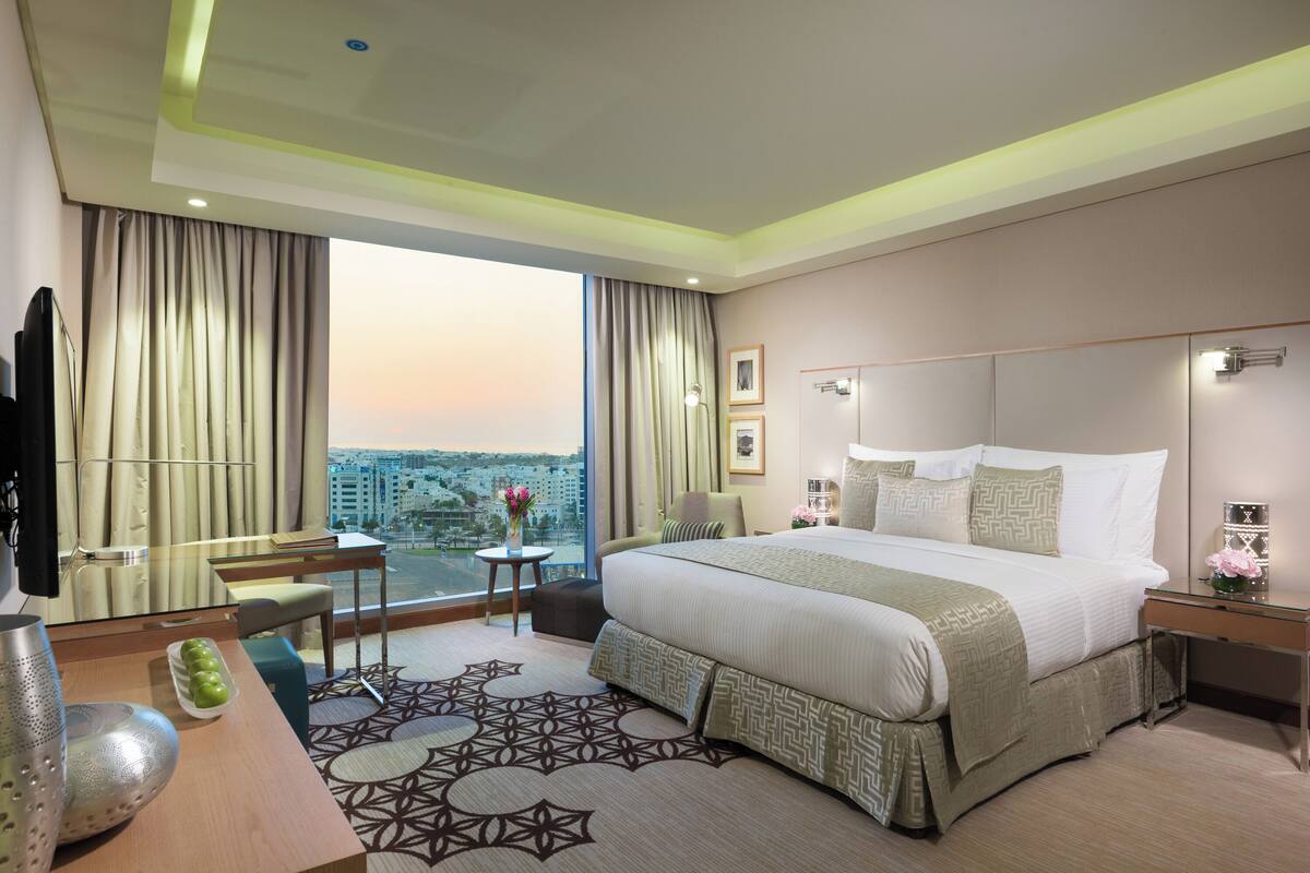 A hotel room with a large bed, a desk, a chair, a TV, and a view of the city.