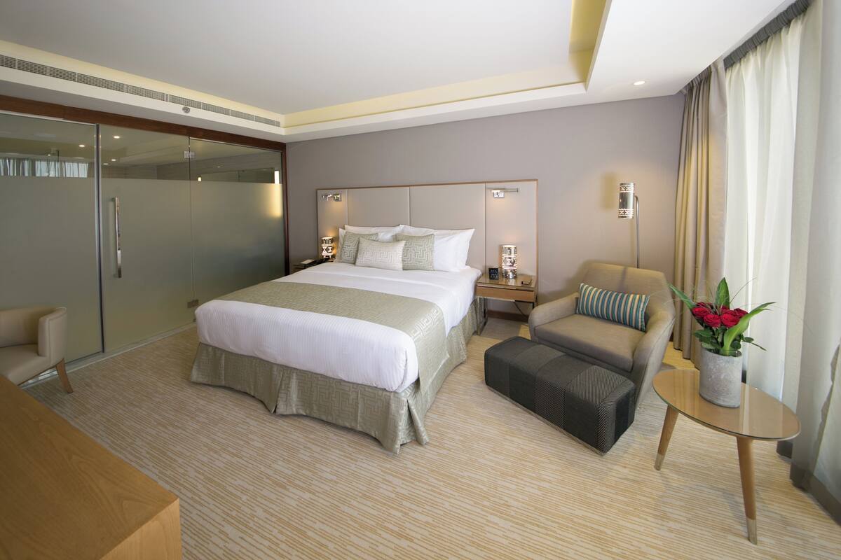 A modern hotel room with a large bed, a sofa, a small table with a vase of flowers, and a glass partition.