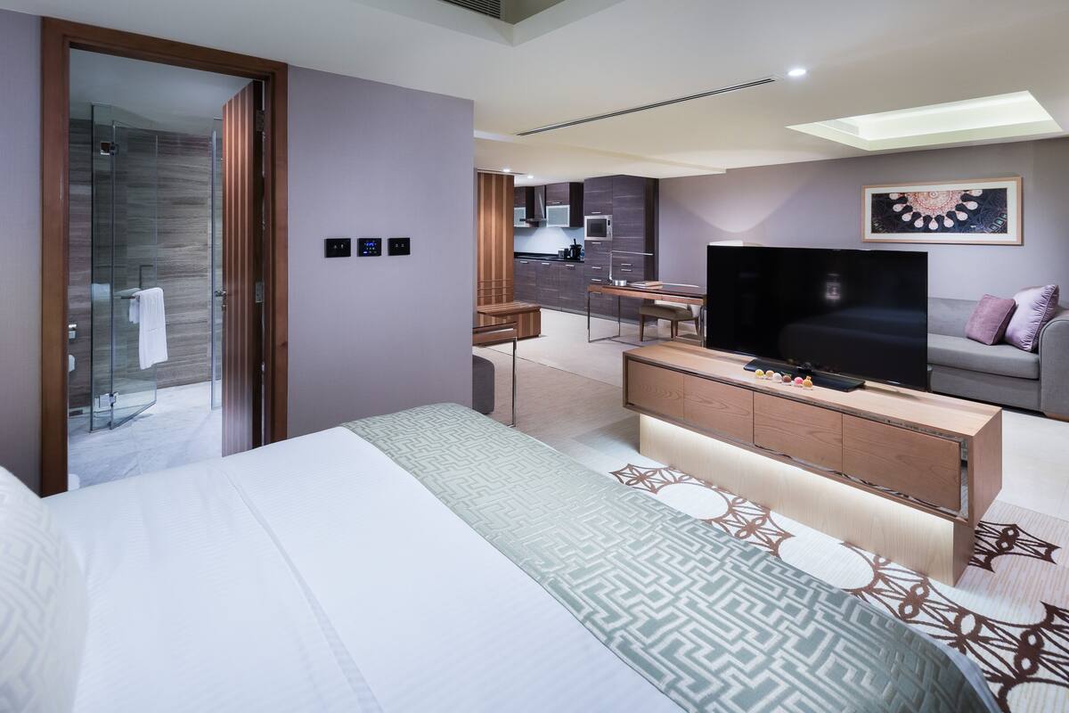 A modern hotel room with a large bed, a TV, a desk, and a glass shower enclosure.