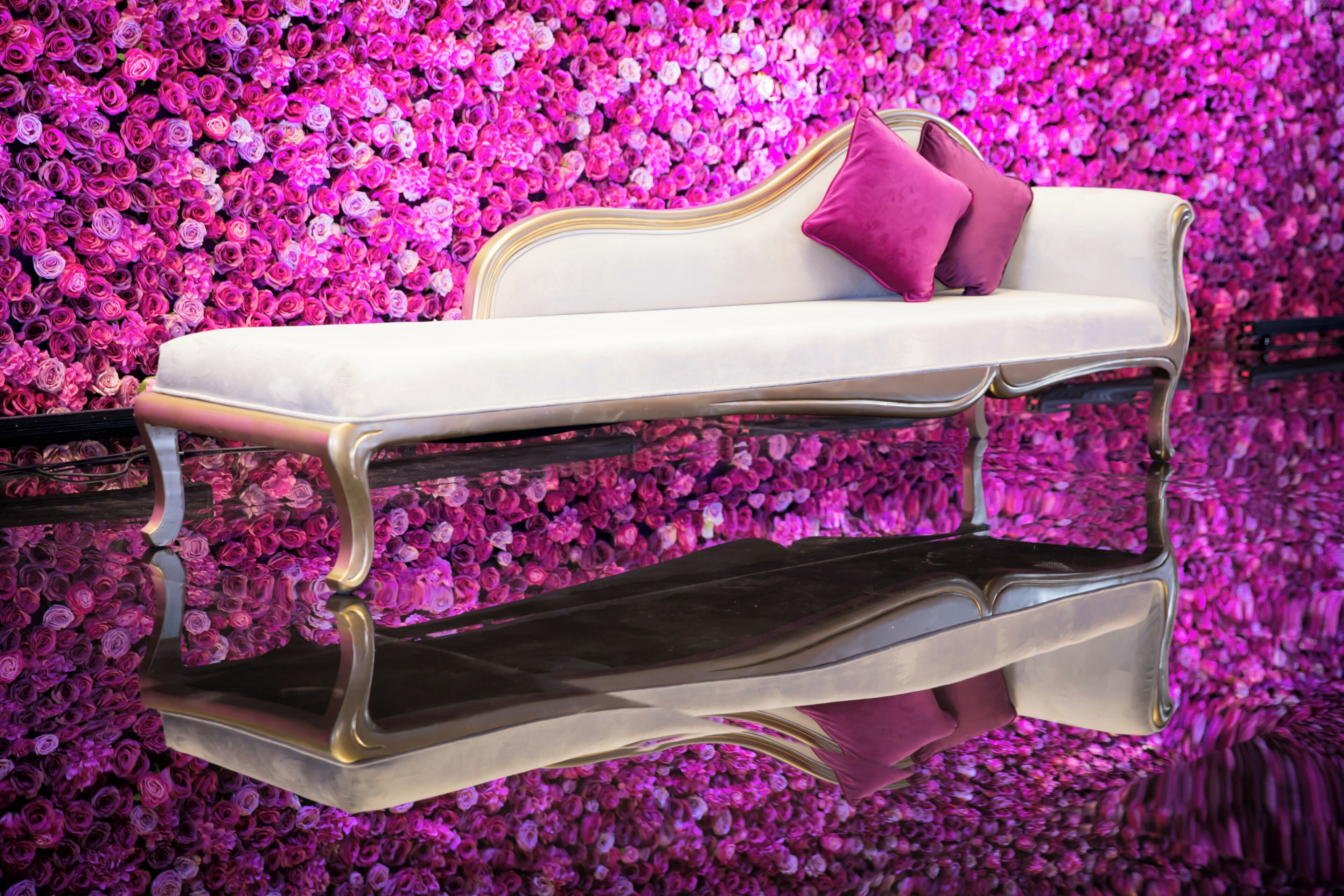 Un sofá blanco con superficie reflectante y una única almohada rosa sobre un fondo de flores rosadas.