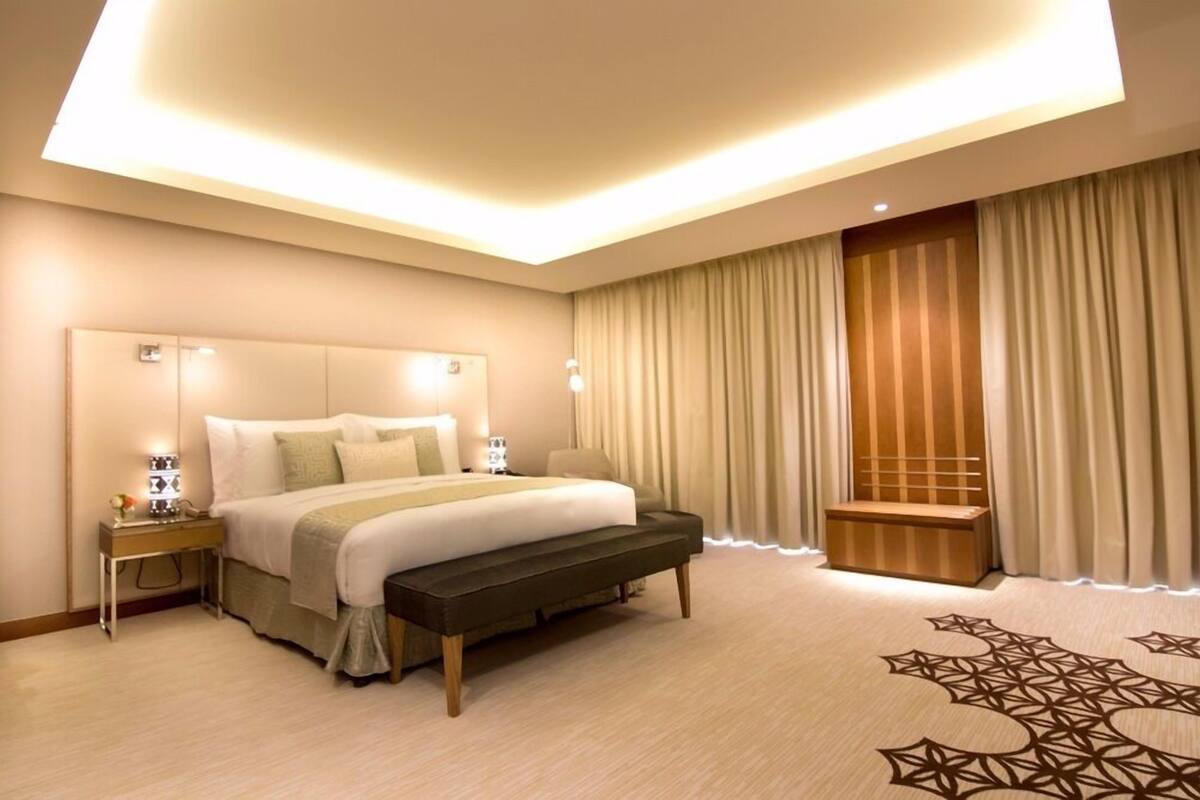 A hotel room with a large bed, bedside lamps, a bench, a nightstand, and a view of a building through the window.